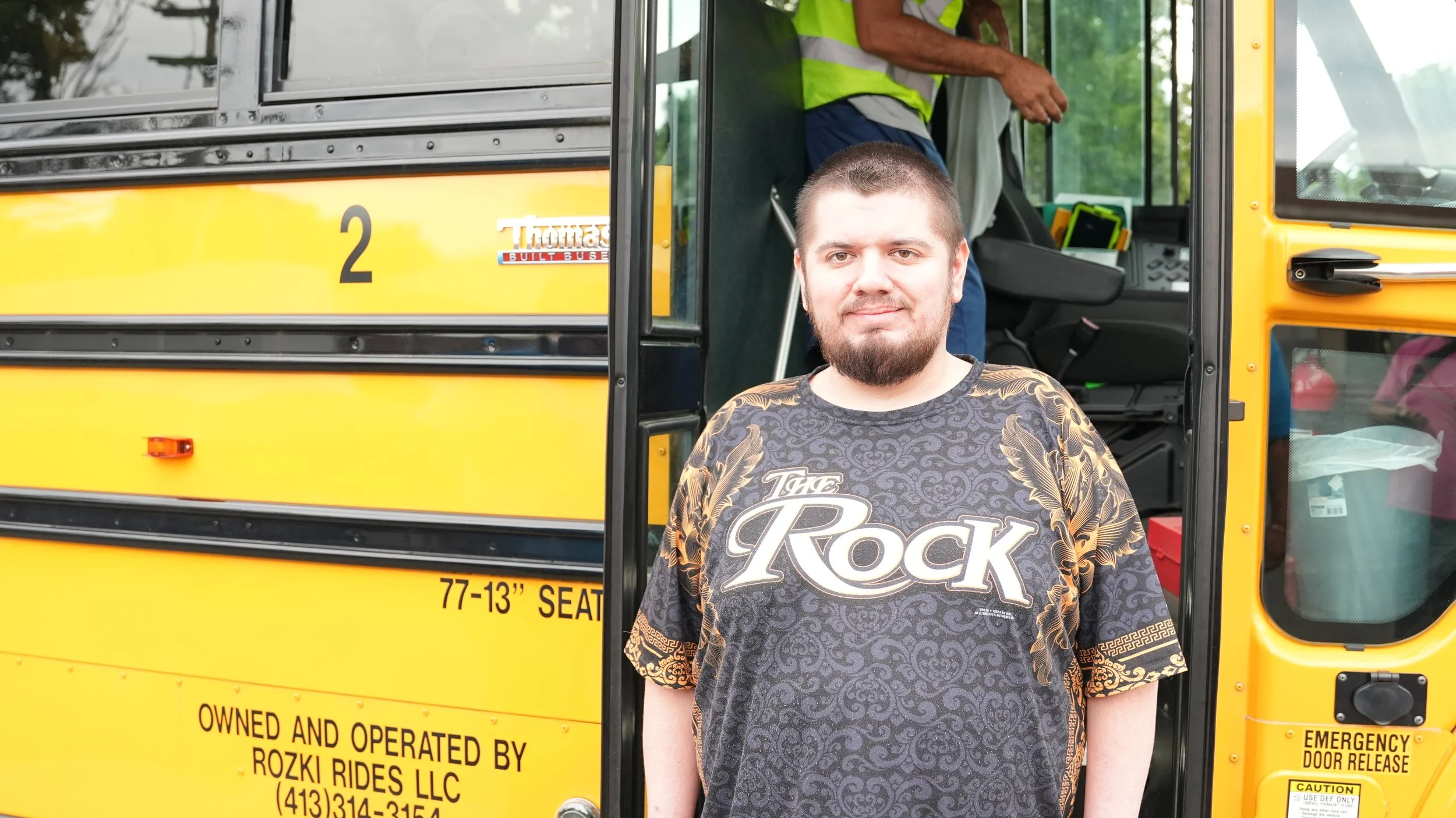 Man standing in front of a yellow school bus with an open door, wearing a black T-shirt with "The Rock" written on it.