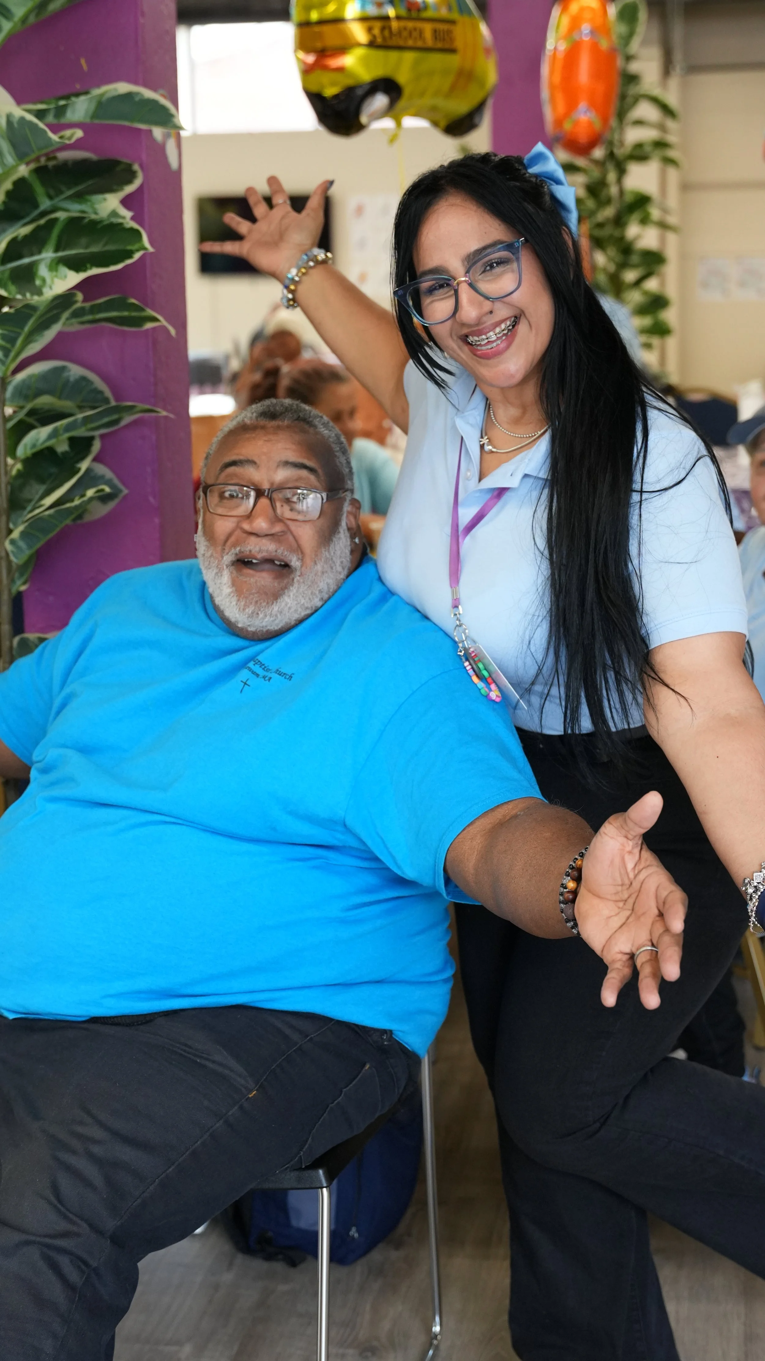 A cheerful woman with long black hair, glasses, and colorful jewelry standing next to a seated man with a gray beard, glasses, and wearing a bright blue shirt. They are in a lively indoor setting, smiling and posing together.