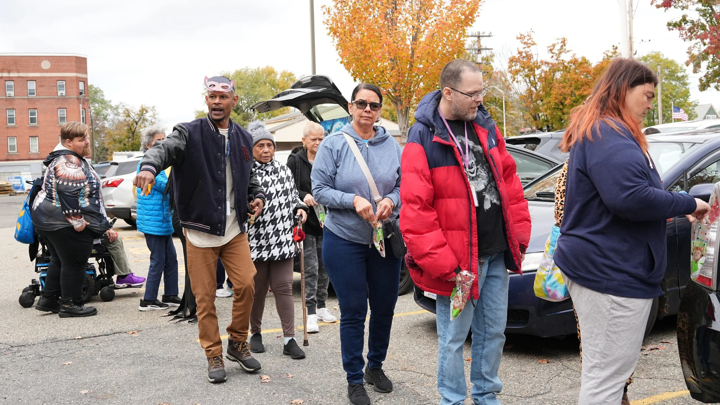 People standing in line outdoors in a parking lot, with some holding holiday-themed items, during fall with colorful trees in the background.