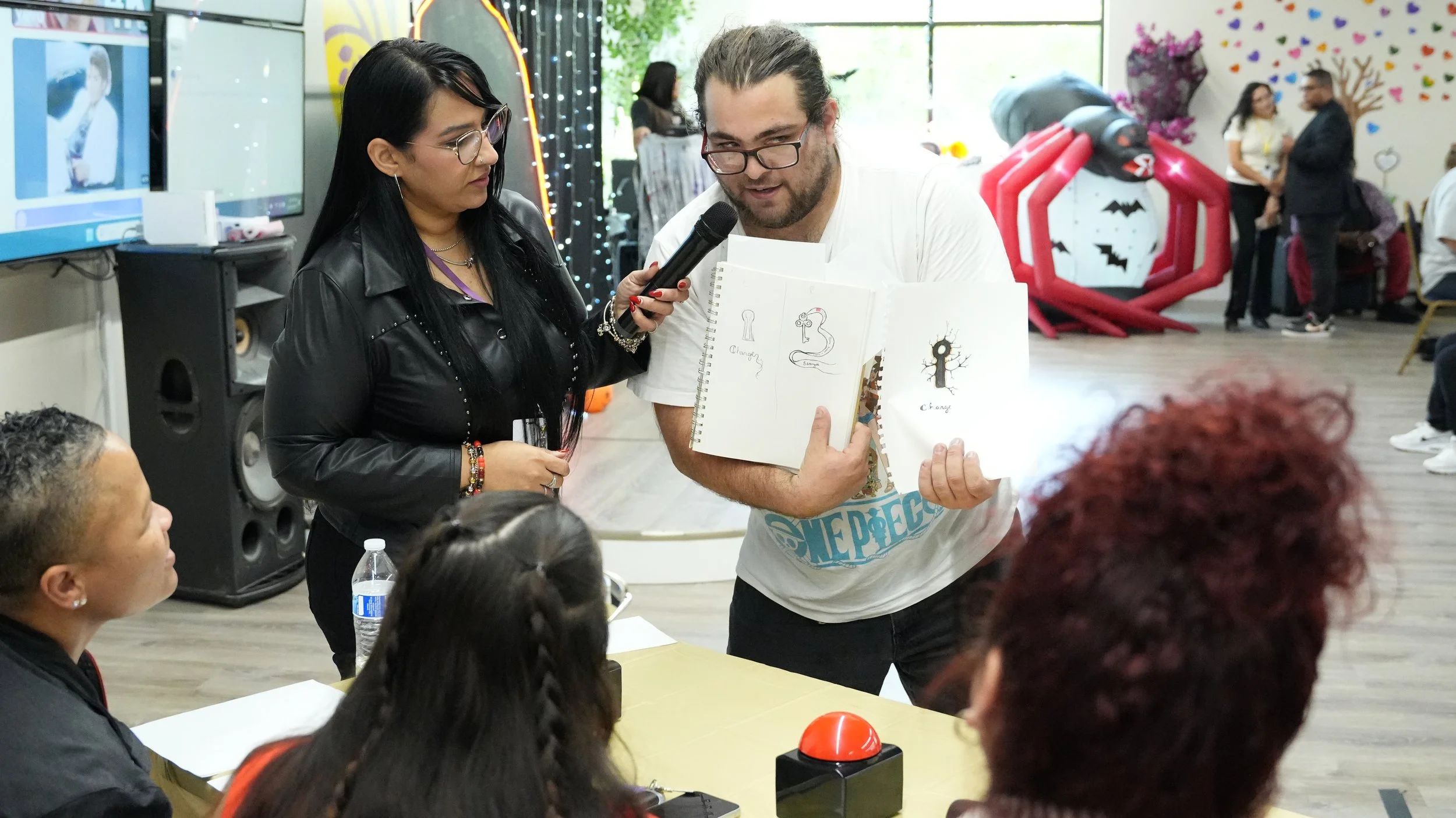 A group of people gathered around a table at an indoor event. A man with glasses is presenting drawings of a snake and a tree, while a woman holds a microphone up to him. Other attendees watch, and a large red inflatable spider decoration, colorful w