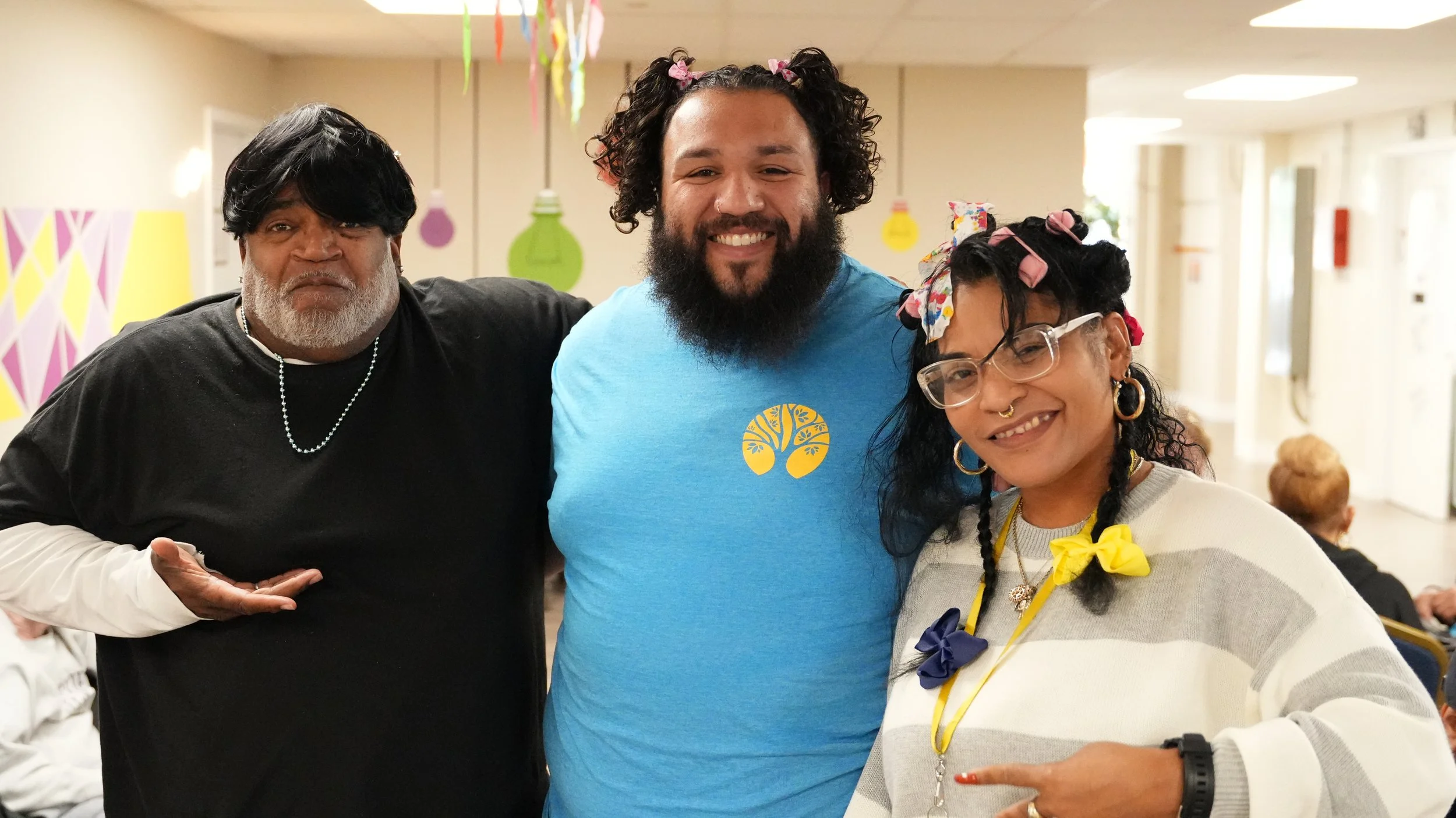 Three people posing together indoors with colorful decorations in the background. They are smiling and appear happy.