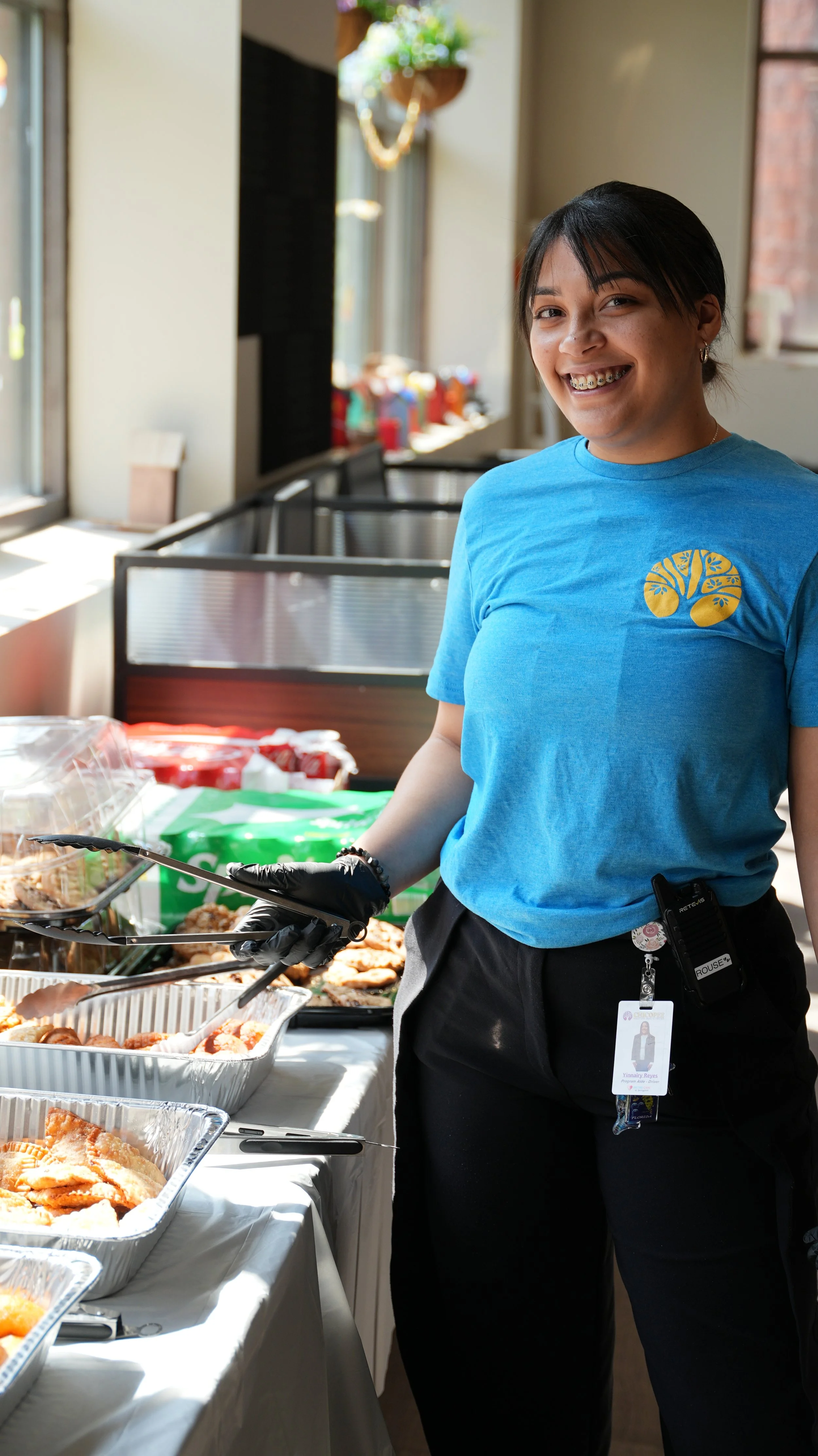 A smiling woman in a blue T-shirt with a tree logo, wearing black gloves and a badge, is standing behind a table with food trays during a food service event.