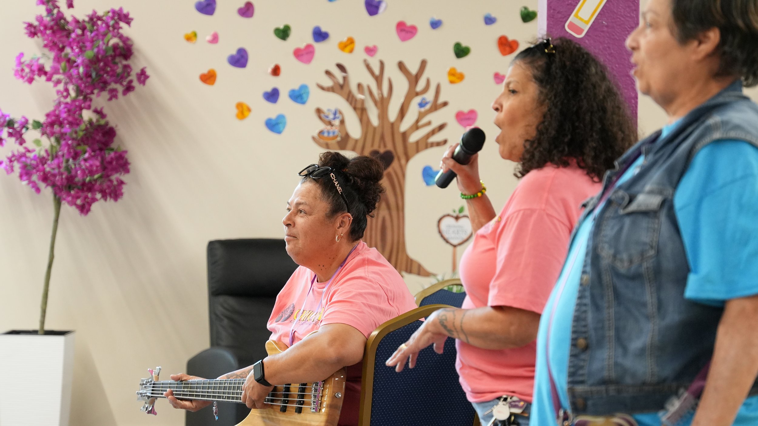 Three women are participating in a musical activity in a decorated room. One woman is playing a guitar, another is singing into a microphone, and the third is observing. Brightly colored heart and puzzle-piece decorations are on the wall, along with 