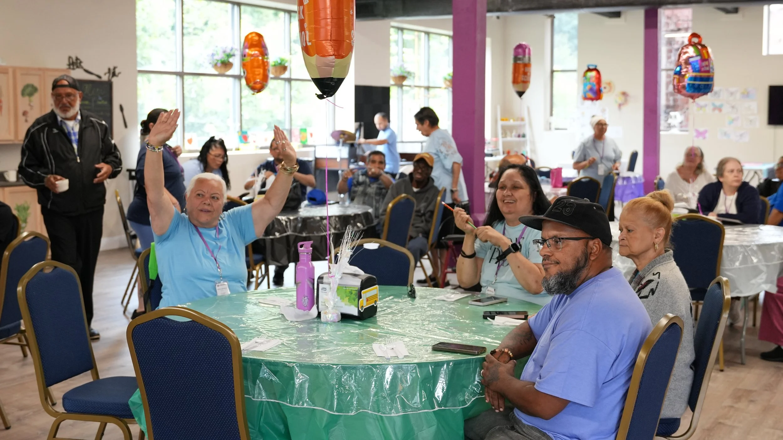 People gathered around a decorated table at a celebration in a large, well-lit room. Some are seated, others are standing, with balloons and colorful decorations hanging from the ceiling. A woman at the table raises her hands, smiling, while others a