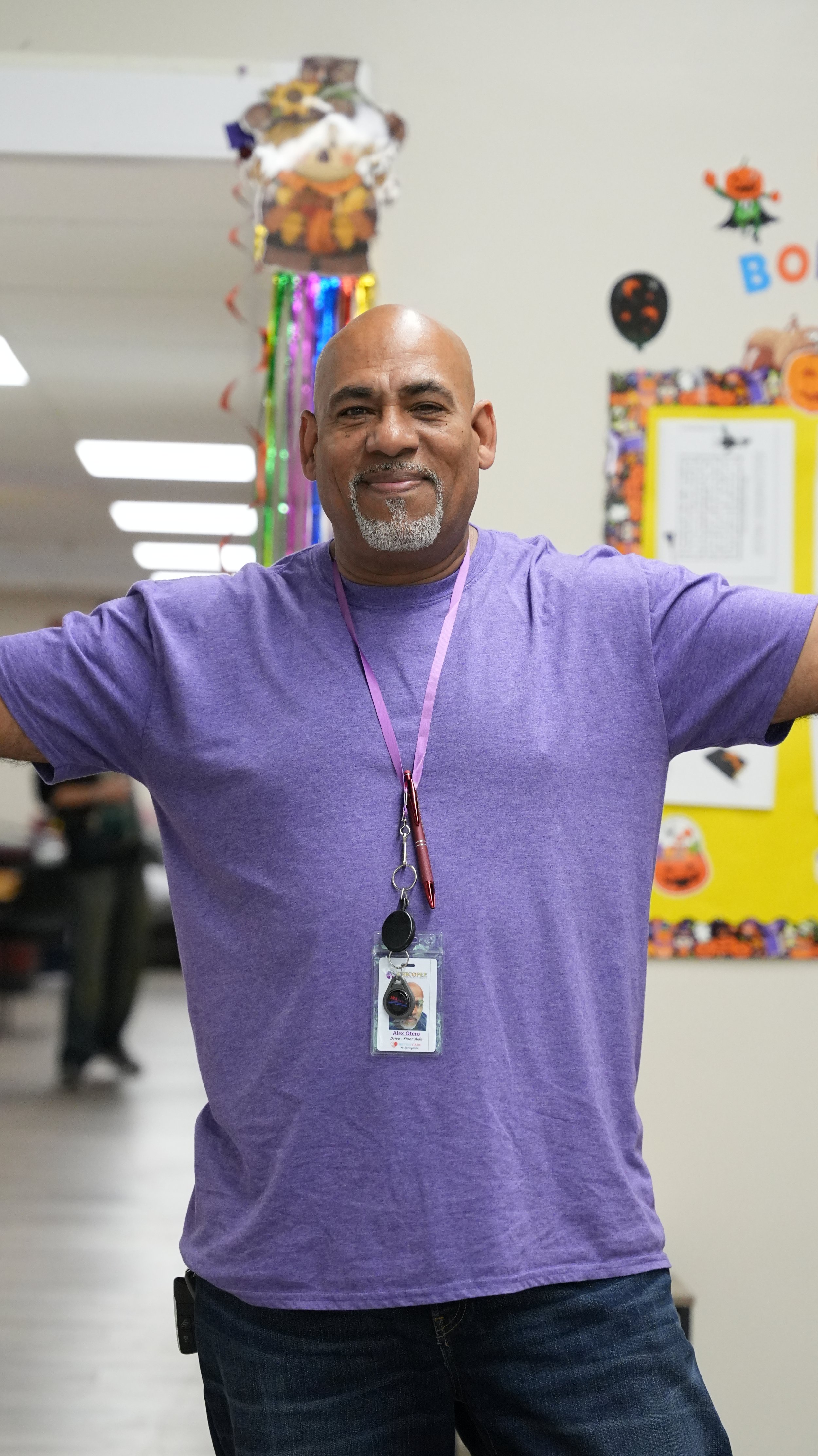 A man in a purple shirt standing in a classroom, smiling with arms outstretched, with Halloween decorations in the background.