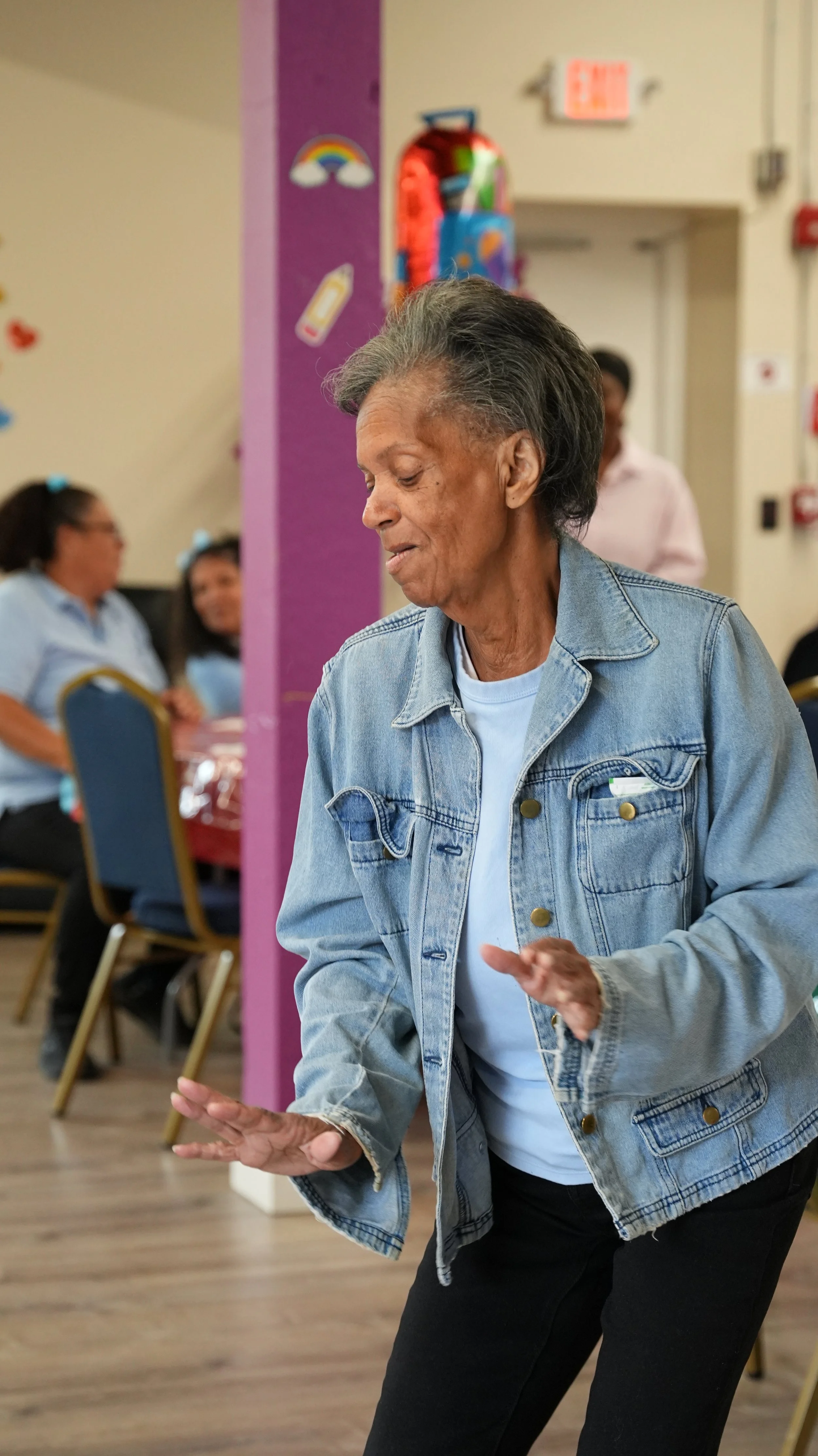 An elderly woman in a denim jacket dancing at a social gathering, with other people sitting at tables in the background.