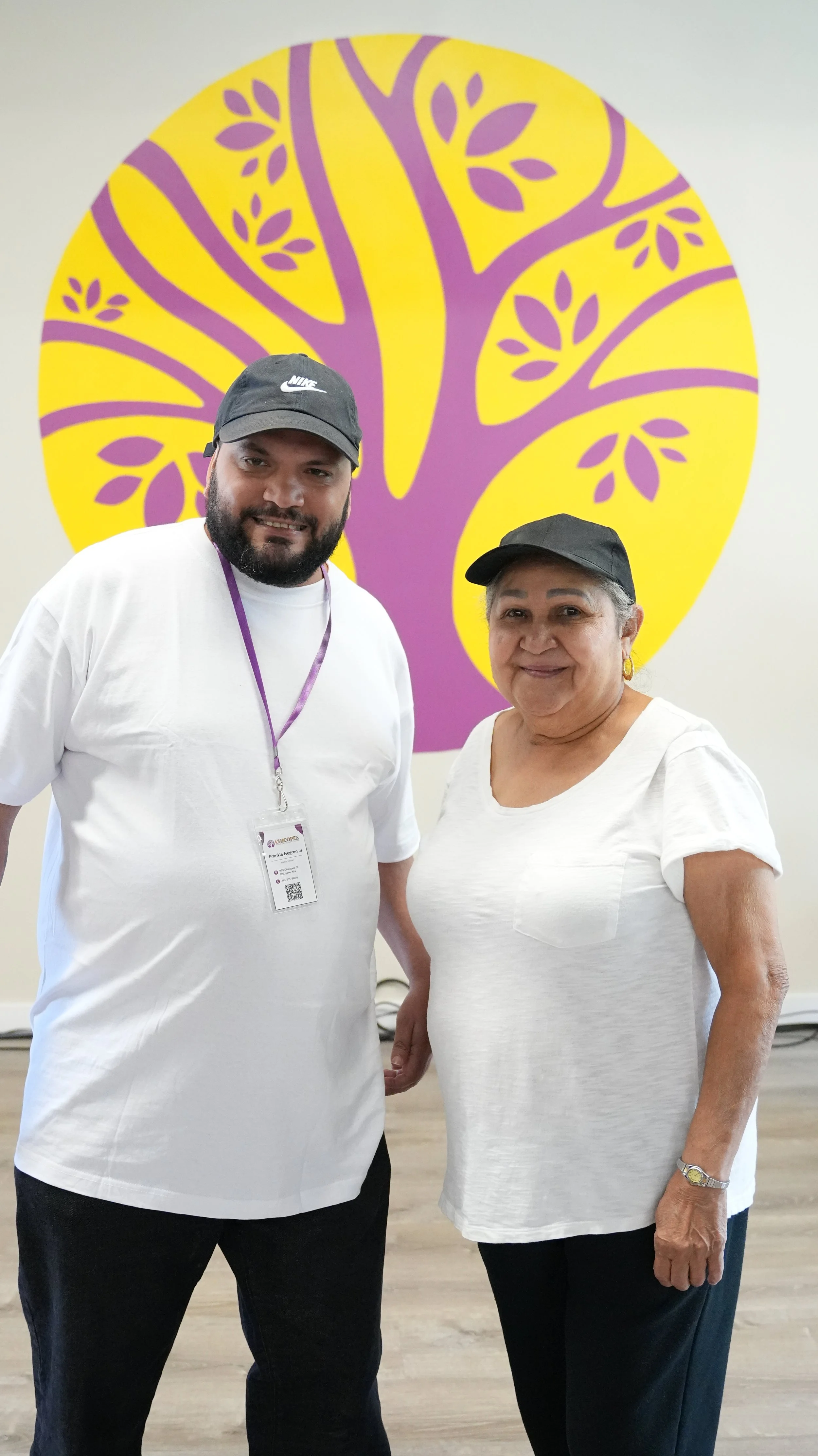 A man and a woman standing in front of a wall with a large purple and yellow tree graphic. Both are wearing white shirts and black caps, smiling at the camera.