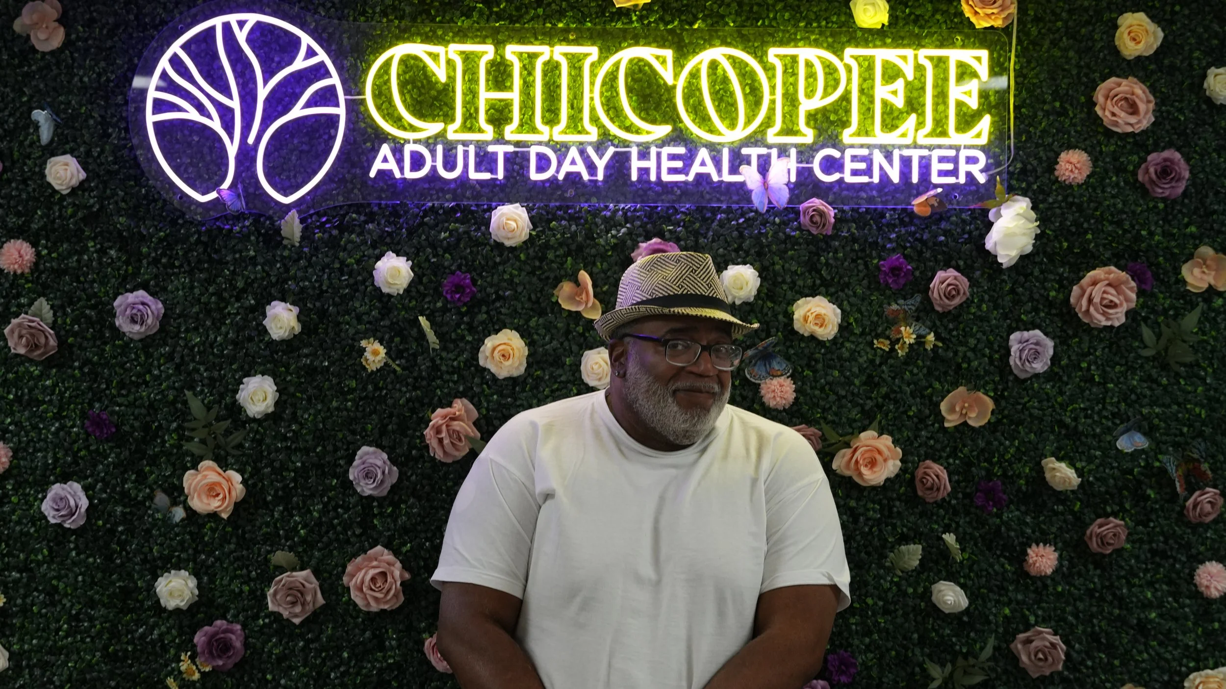 A man with gray hair, beard, and glasses, wearing a white t-shirt and a straw fedora with a black band, standing in front of a floral wall with a neon sign reading "Chicopée Adult Day Health Center" and neon tree logo.