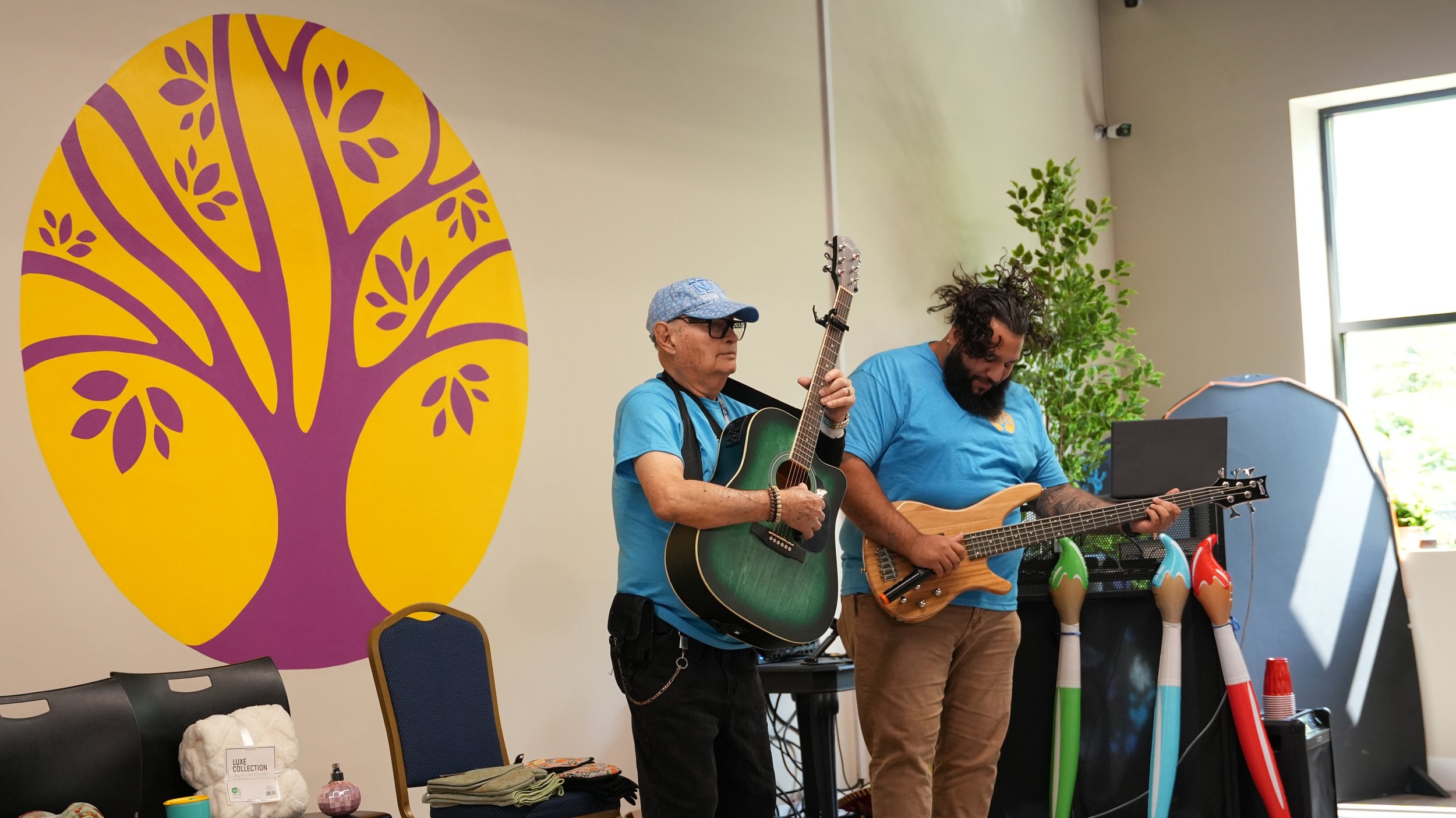 Two men playing guitars in a room with a large purple and yellow tree mural on the wall. One man is older with glasses and a blue cap, holding an acoustic guitar. The other is younger, with curly hair and a beard, playing an electric guitar. There ar