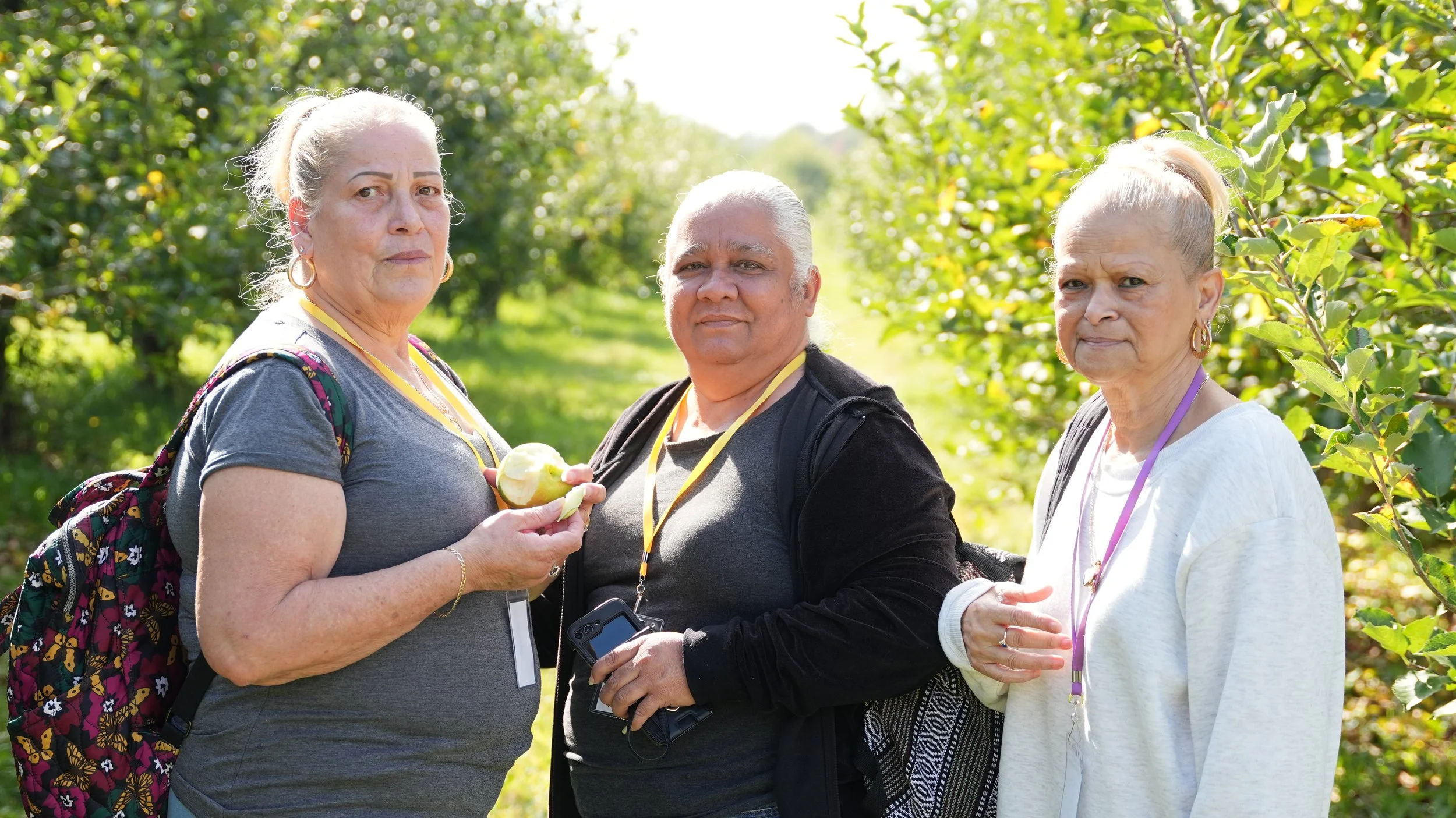 Three older adults standing in a lush orchard, two women and one man, holding a partially eaten apple, wearing badges on lanyards, with trees and sunlight in the background.