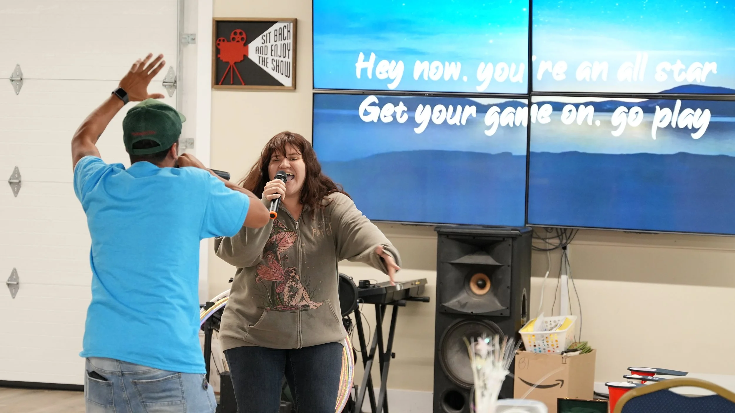 A woman and a man singing and dancing together at a karaoke night, with a large screen displaying lyrics behind them.