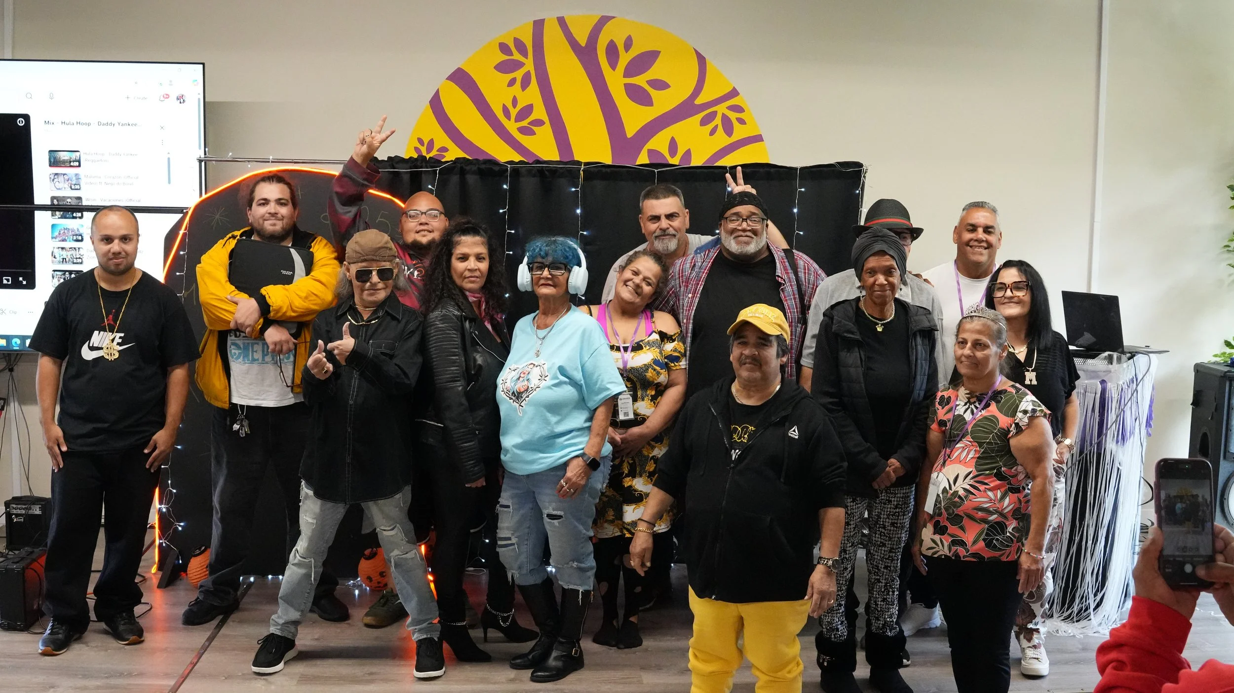 A group of people posing indoors in front of a decorated backdrop with purple and yellow artwork and string lights. Some are smiling, making peace signs, or standing casually.