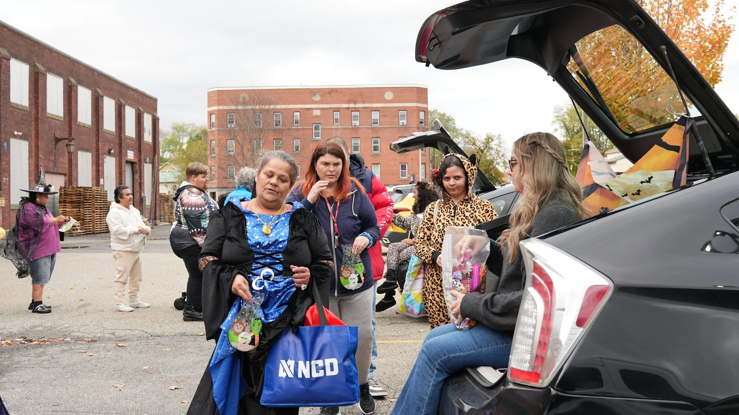 People gathered outdoors dressed in costumes for Halloween, including a woman in a blue dress with a cape, another woman in a leopard print hoodie, and individuals in witch costumes, next to open car trunks decorated for the occasion.