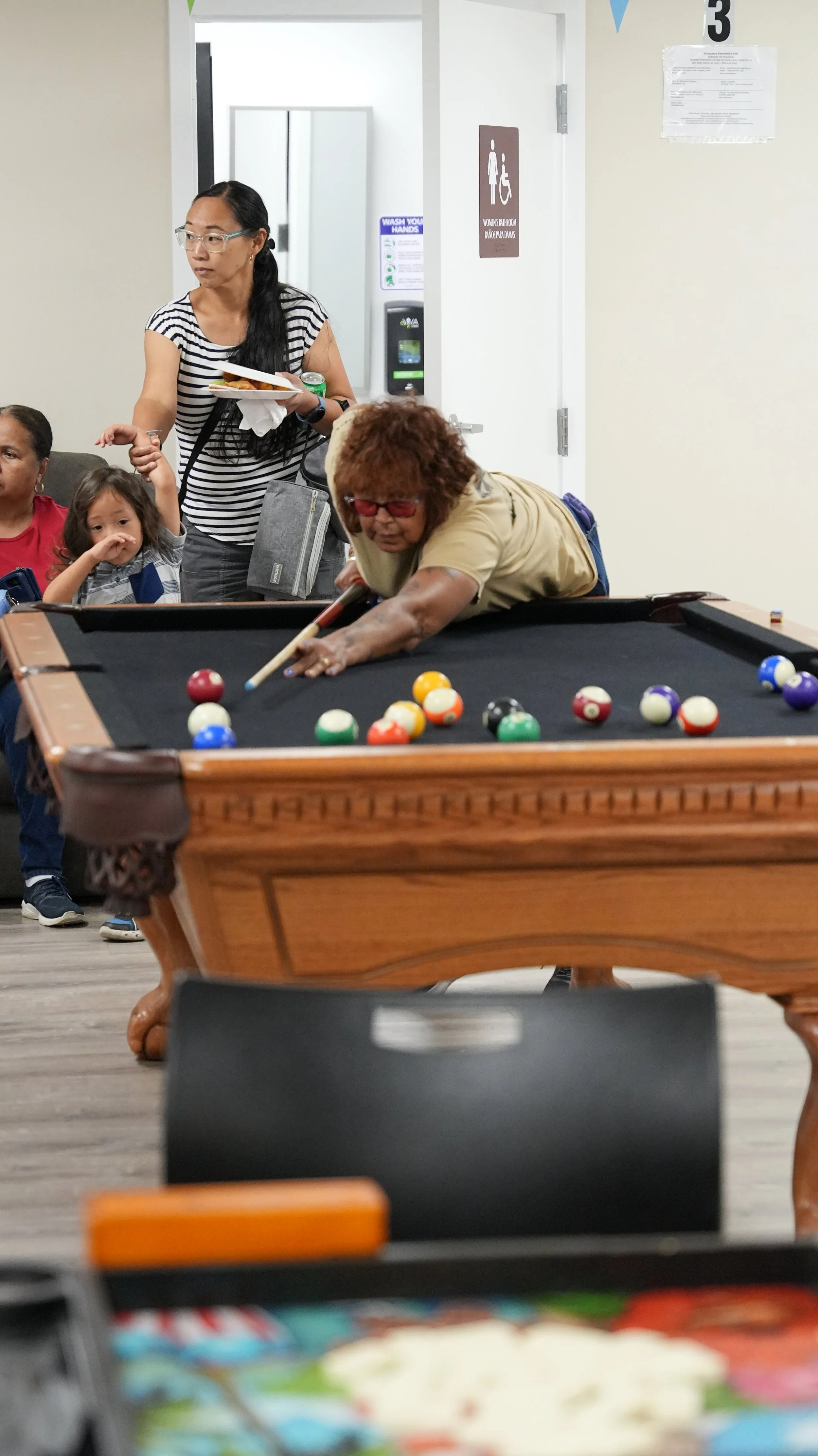 A woman playing pool at a social gathering while several children and another woman watch nearby. The scene is indoors, in a recreational or community room.