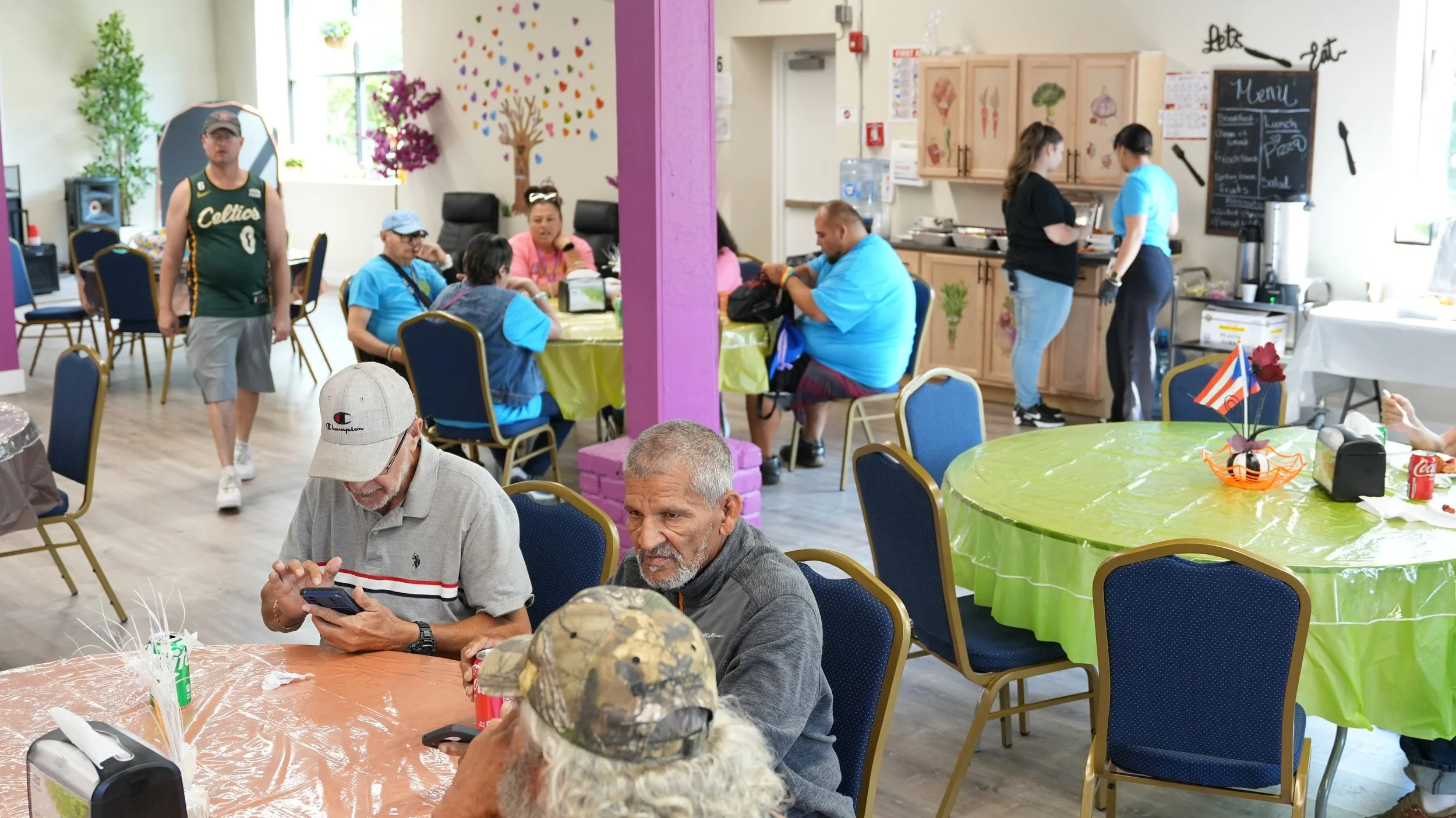 A group of people, mostly elderly, sitting at tables in a community room or cafeteria, with some using their phones and others talking. There is a colorful decoration with a tree and hearts on the wall, and a buffet area in the background with two wo
