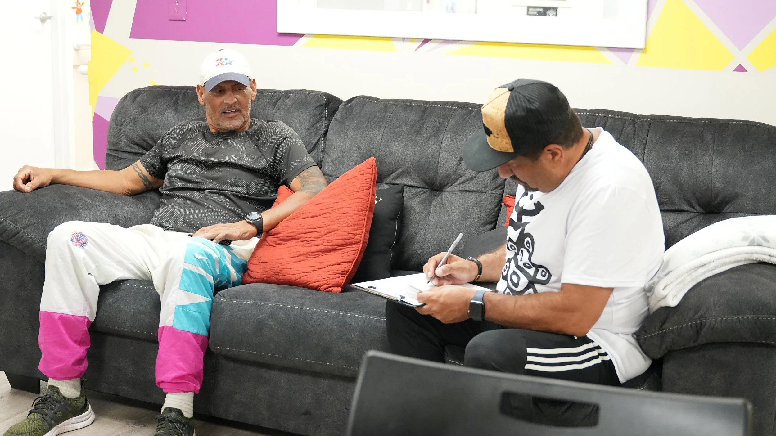 Two men sitting on a dark grey couch, one is writing on a clipboard and the other is looking at him, in a room with colorful geometric wall art.