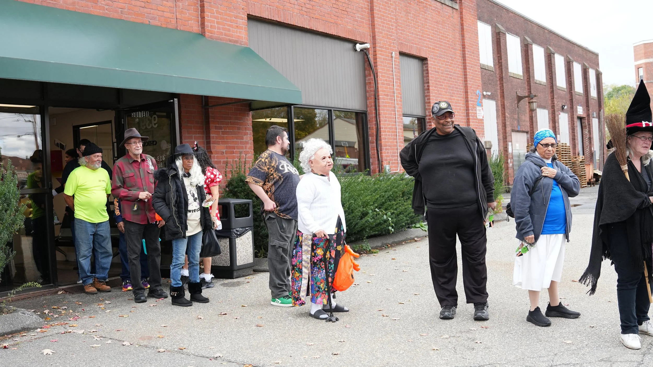 People standing in a line outside a brick building with a green awning, some wearing Halloween costumes and casual clothing.