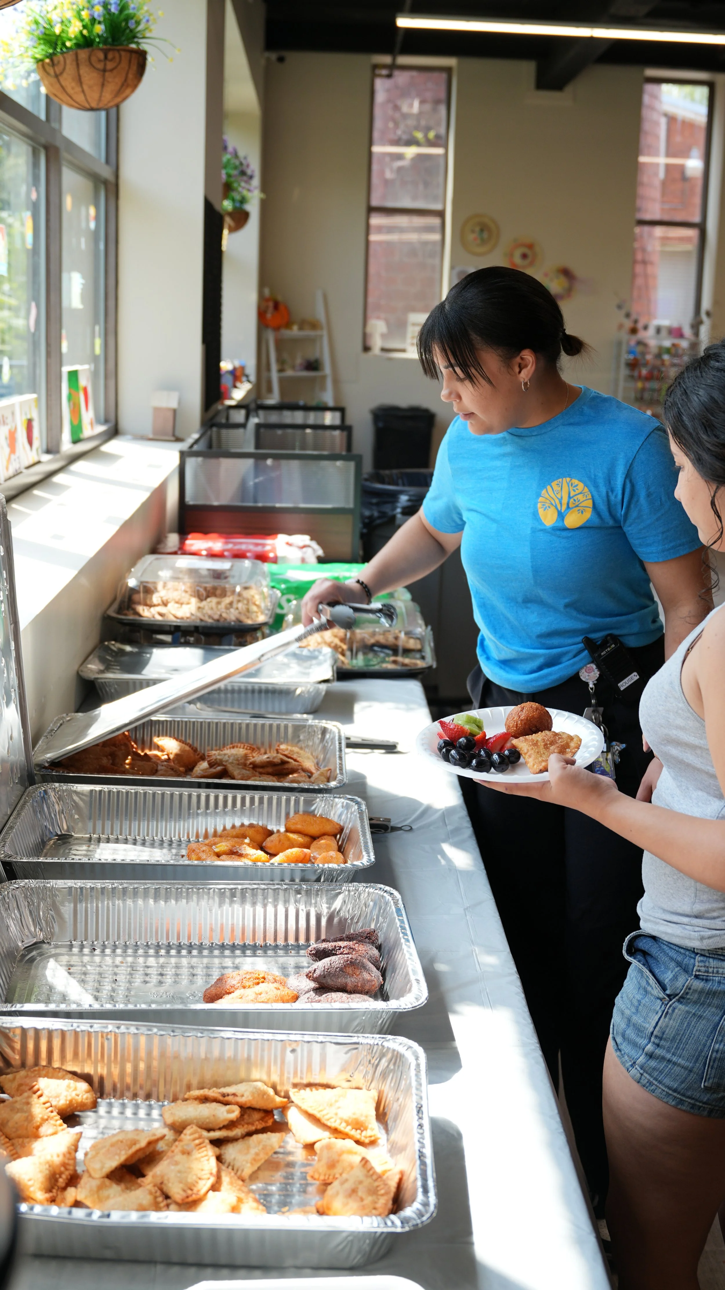 A woman in a blue shirt serving food at a buffet with trays of various dishes, while another woman in a gray tank top is holding a plate of fruit and dessert.