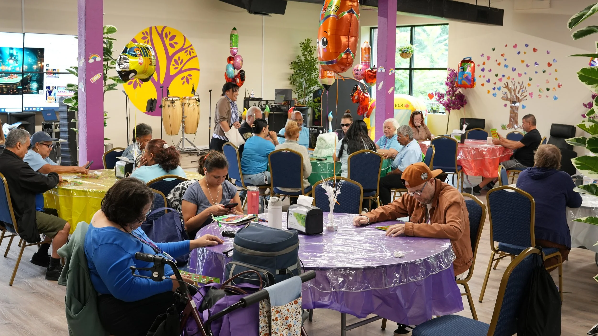 Indoor gathering of people, some with disabilities, at a lively event with colorful balloons, decorated tables, and children’s artwork on walls, during daytime.