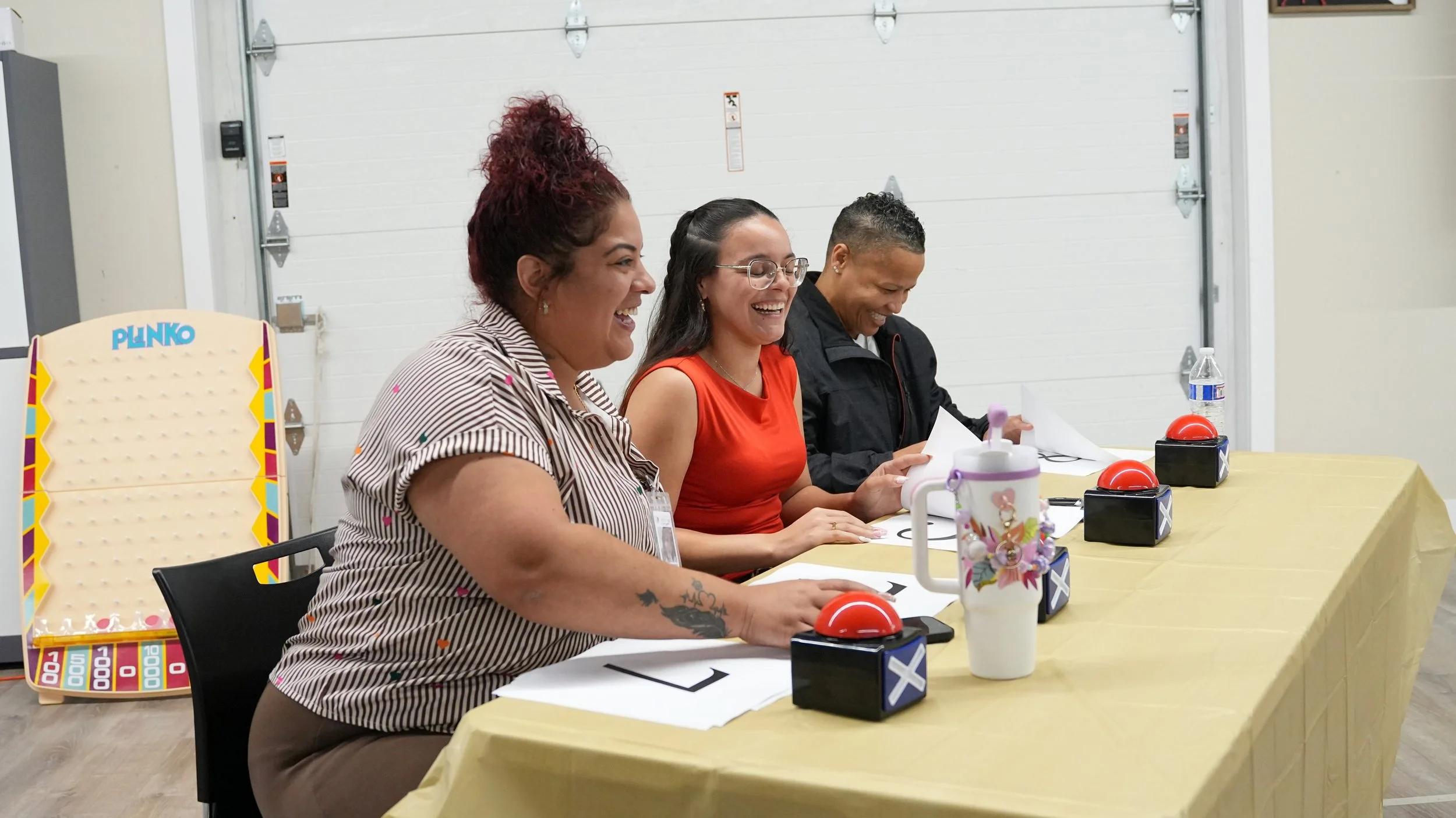 Three women sitting at a table with red buzzer buttons, papers, a water bottle, and a colorful cup, smiling and laughing.