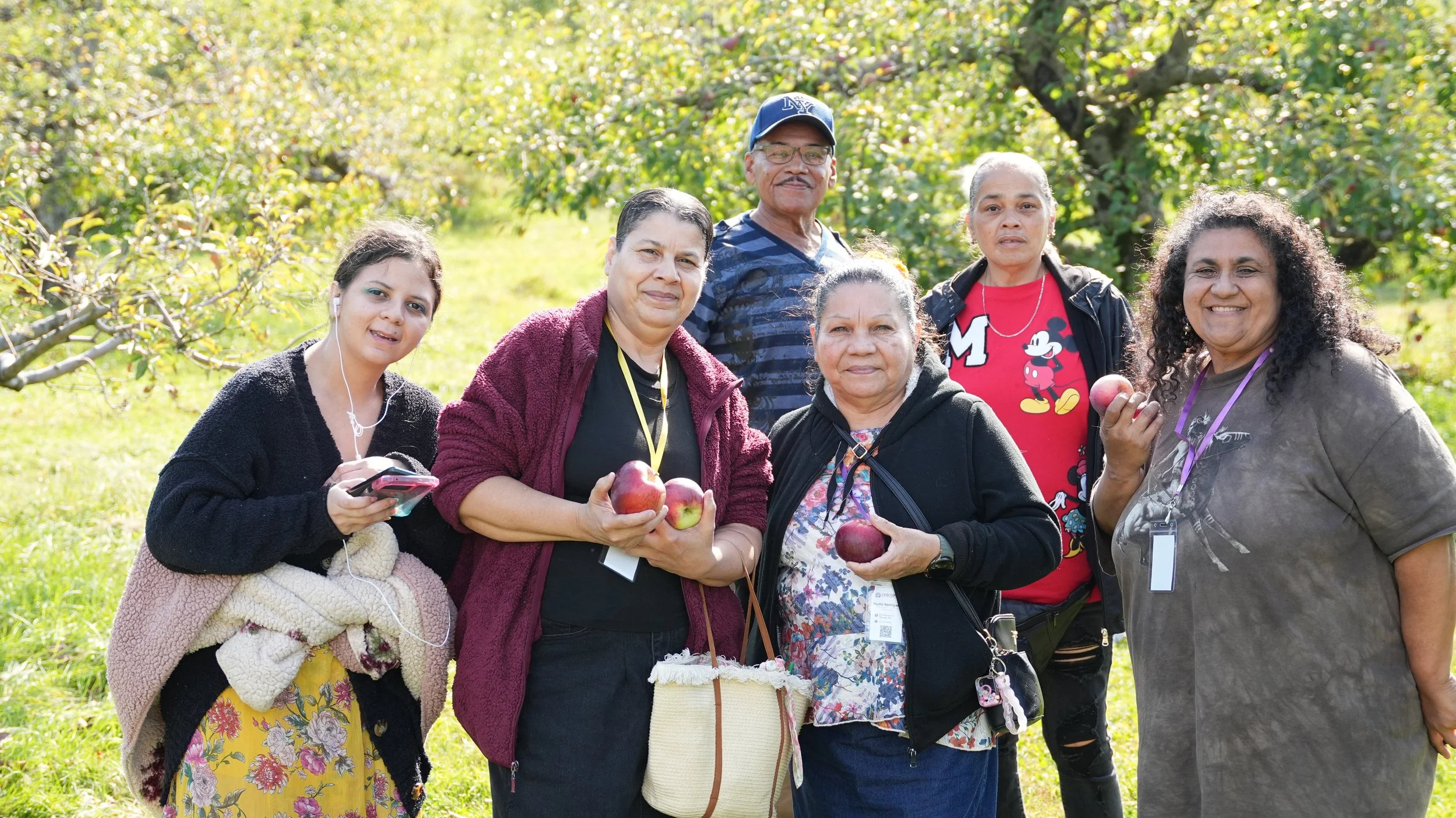 A group of six people standing outdoors in apple orchard, holding apples and smiling, with trees and sunlight in the background.