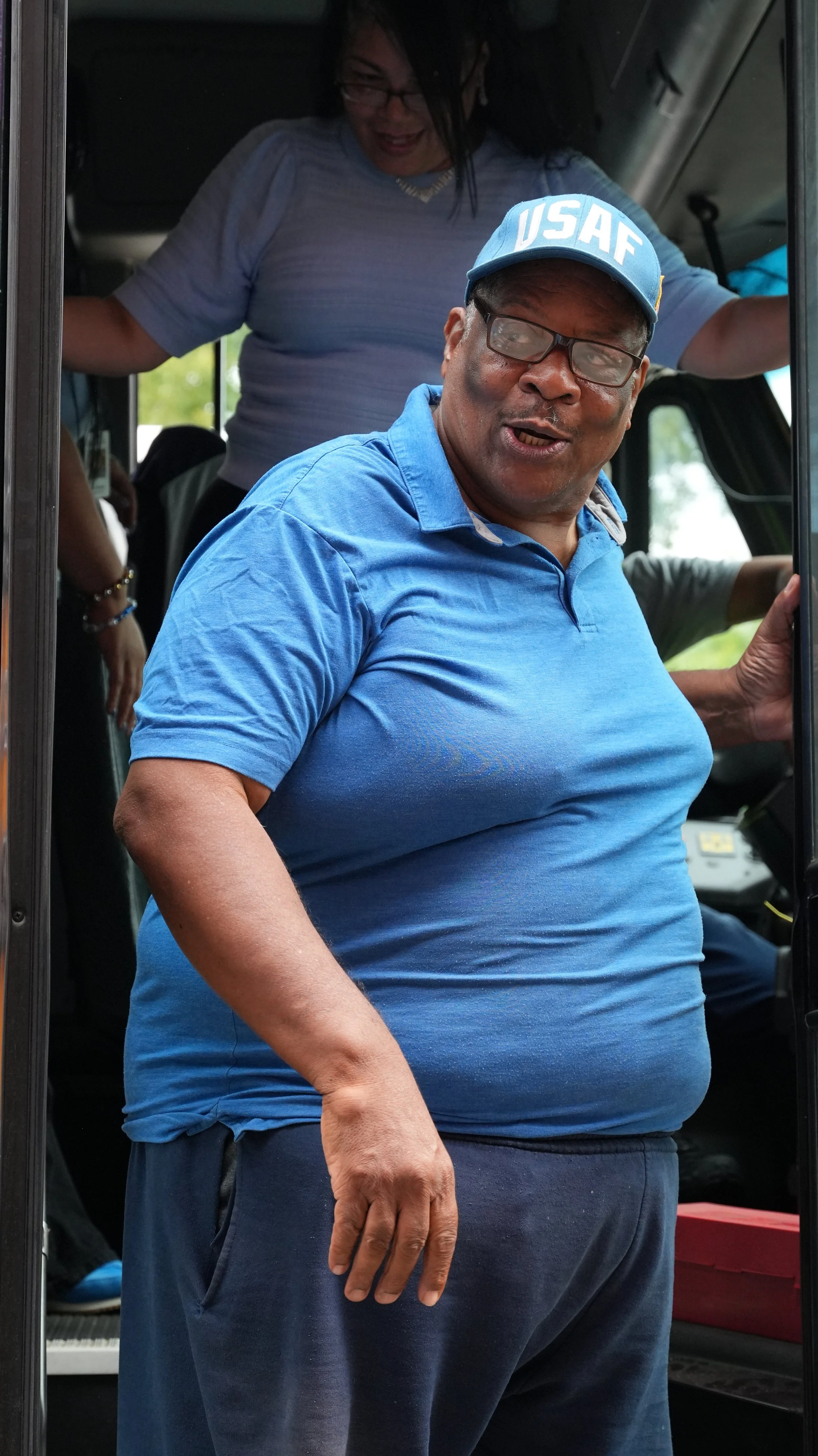 A man wearing glasses, a blue USF cap, and a blue polo shirt standing on a bus or train, with a woman in the background.