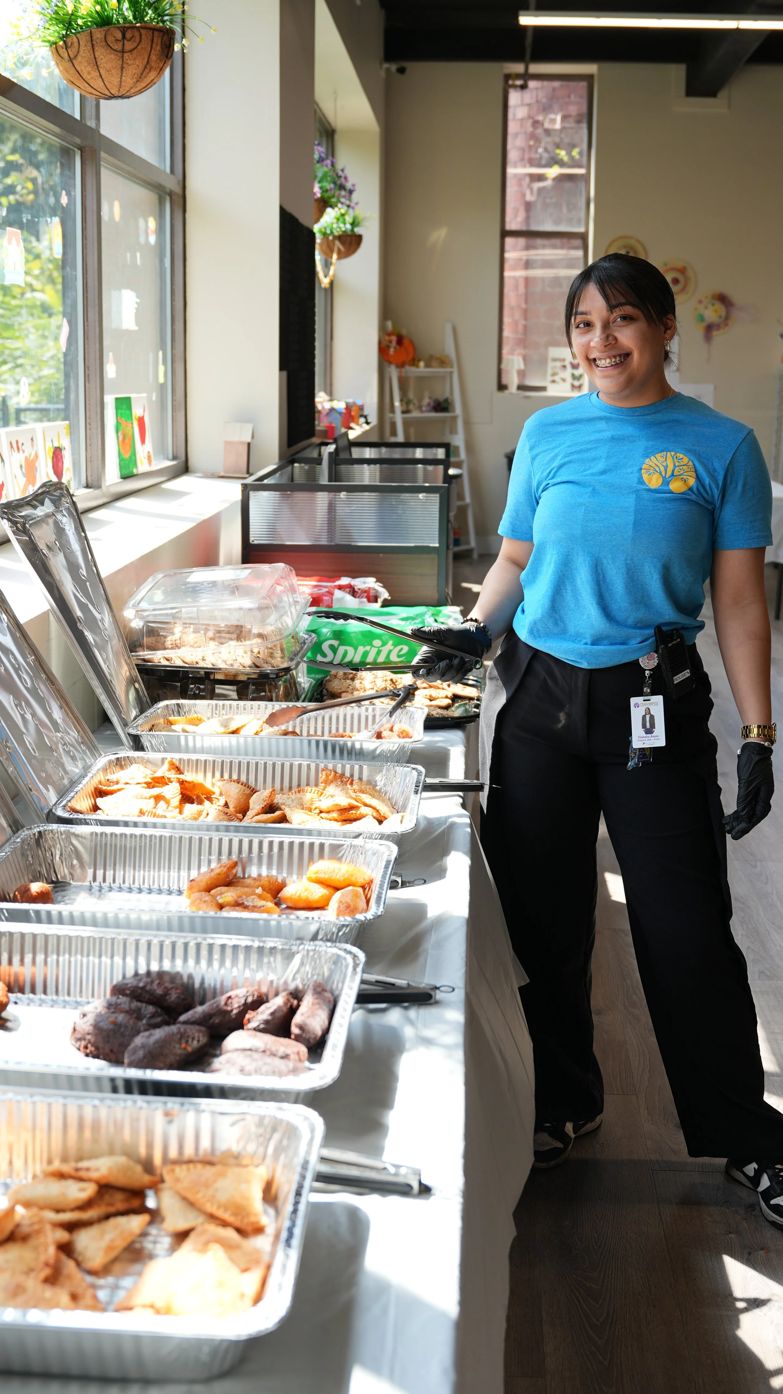 A woman wearing a blue shirt and black pants, with a clip badge and black gloves, stands next to a table with trays of assorted food in a brightly lit room with large windows and hanging baskets of plants.