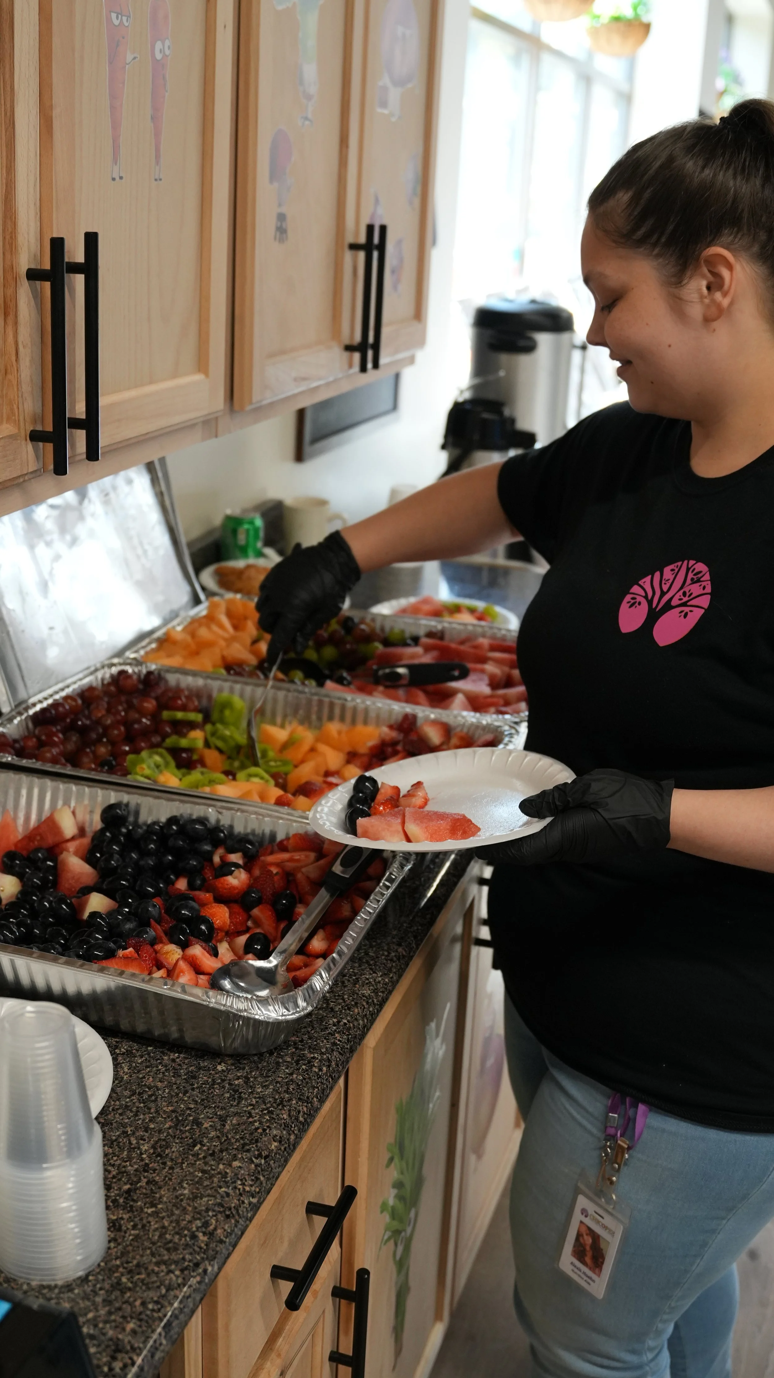 A woman serving herself fruit from a buffet with trays of grapes and sliced melons and berries.
