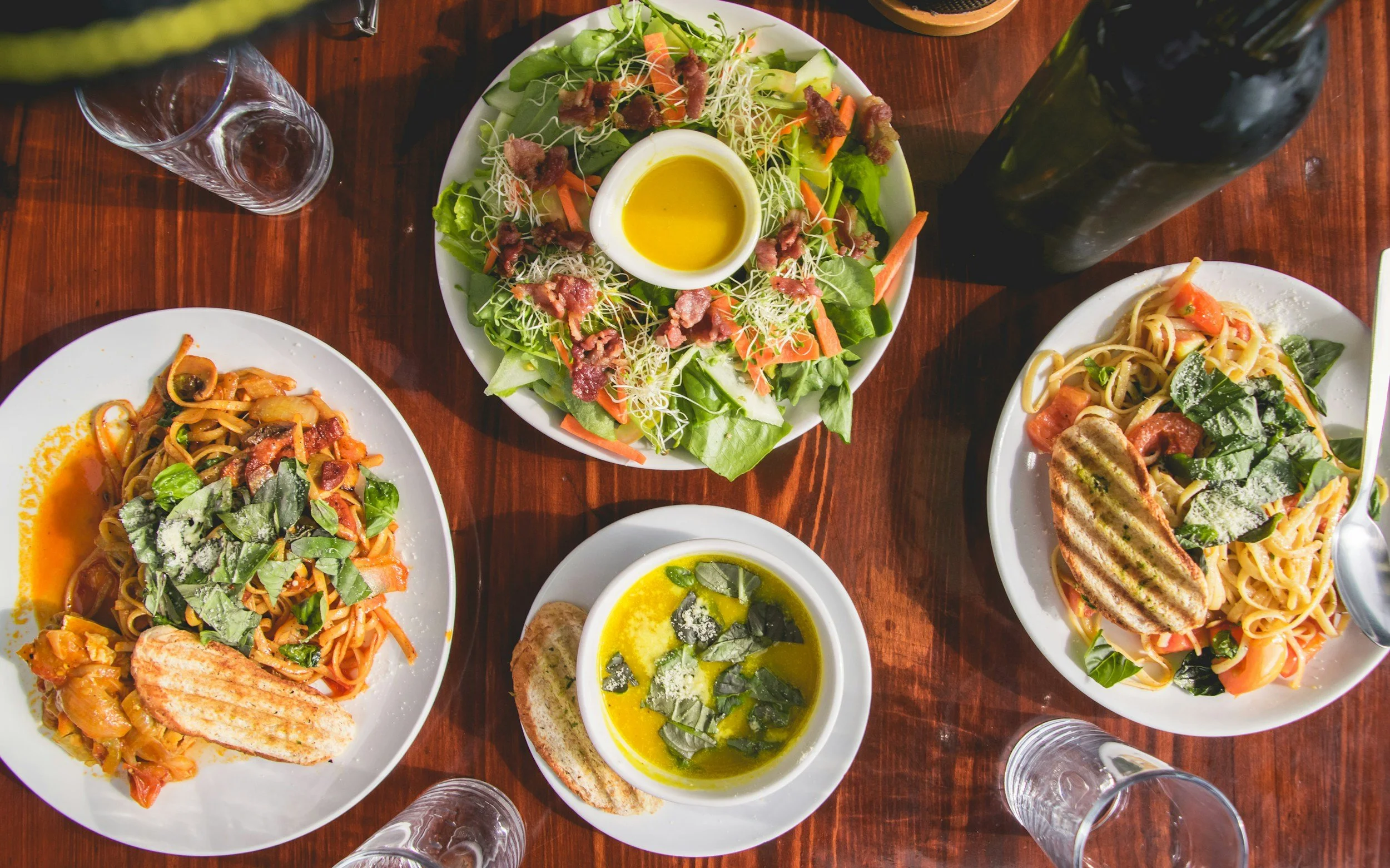 A top-down view of a wooden table with a salad, two plates of pasta, a bowl of soup, and glasses of water.