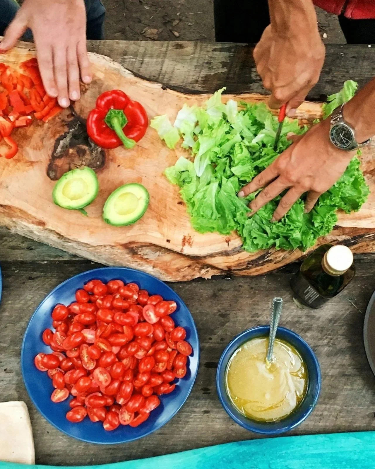 People preparing a fresh vegetable salad with cherry tomatoes, avocado, lettuce, cut bell pepper, and a bowl of dressing on a rustic wooden table.