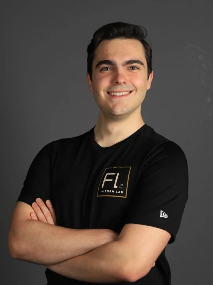 A young man with dark hair and fair skin, smiling with arms crossed, wearing a black t-shirt.