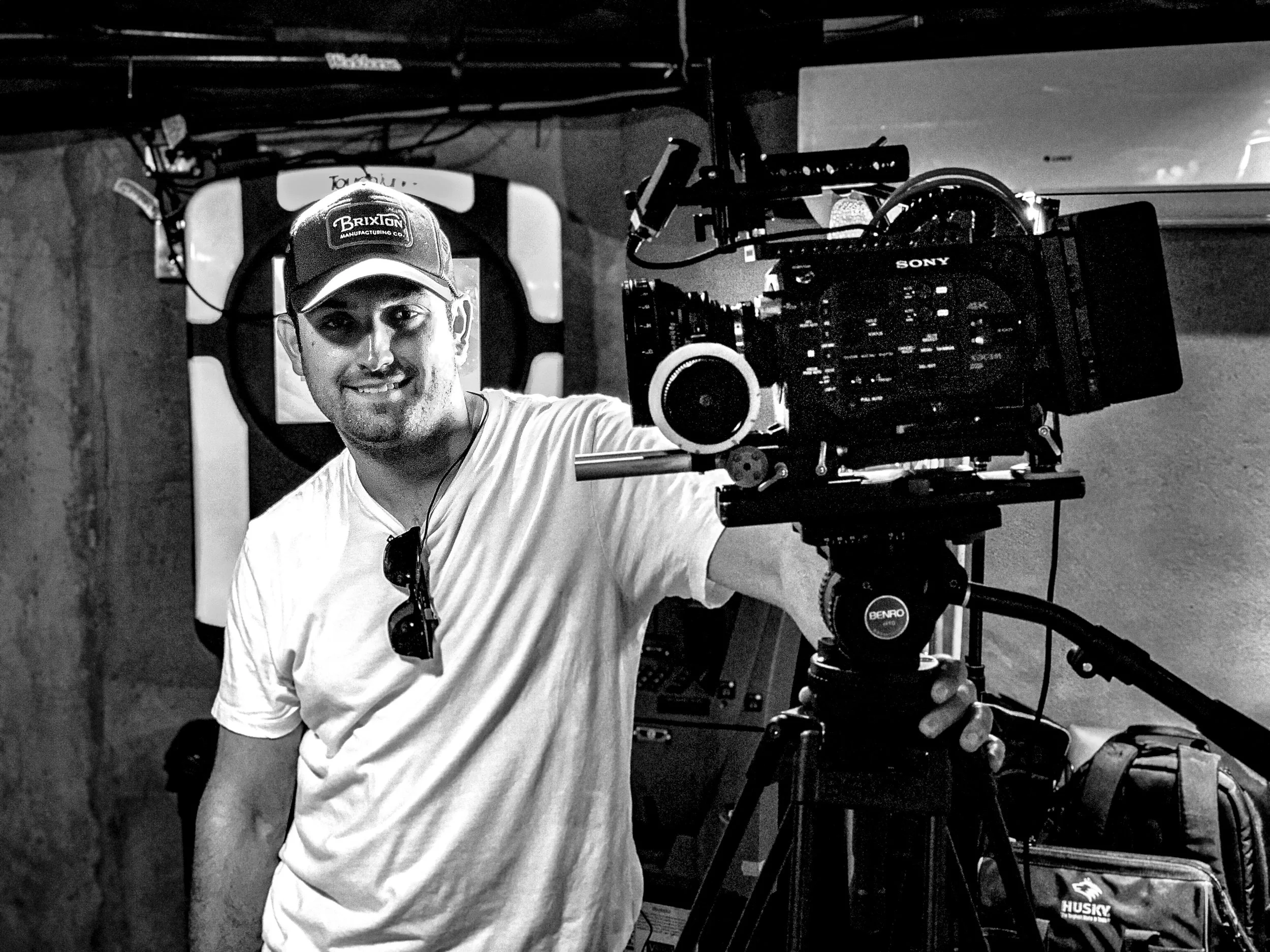 A man with a cap and sunglasses hanging from his shirt operating a professional video camera on a tripod in a workshop or studio.