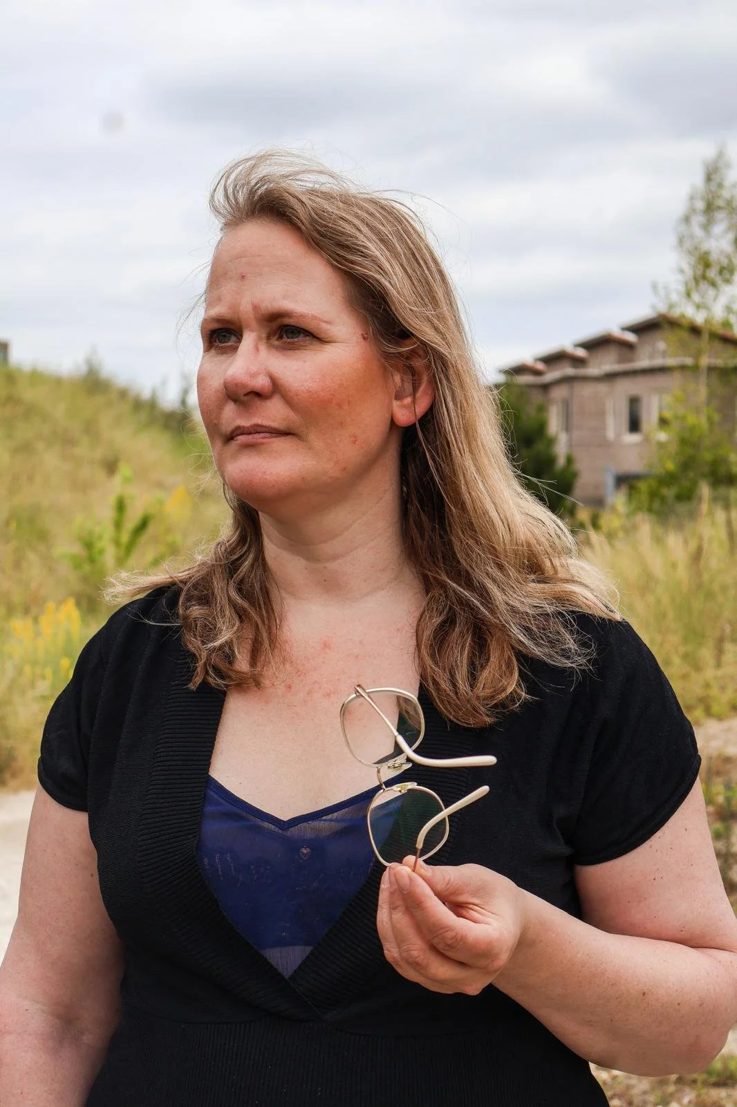 Vrouw met bril in haar hand die buiten staat, met een barren veld en een gebouw op de achtergrond, onder een bewolkte hemel.