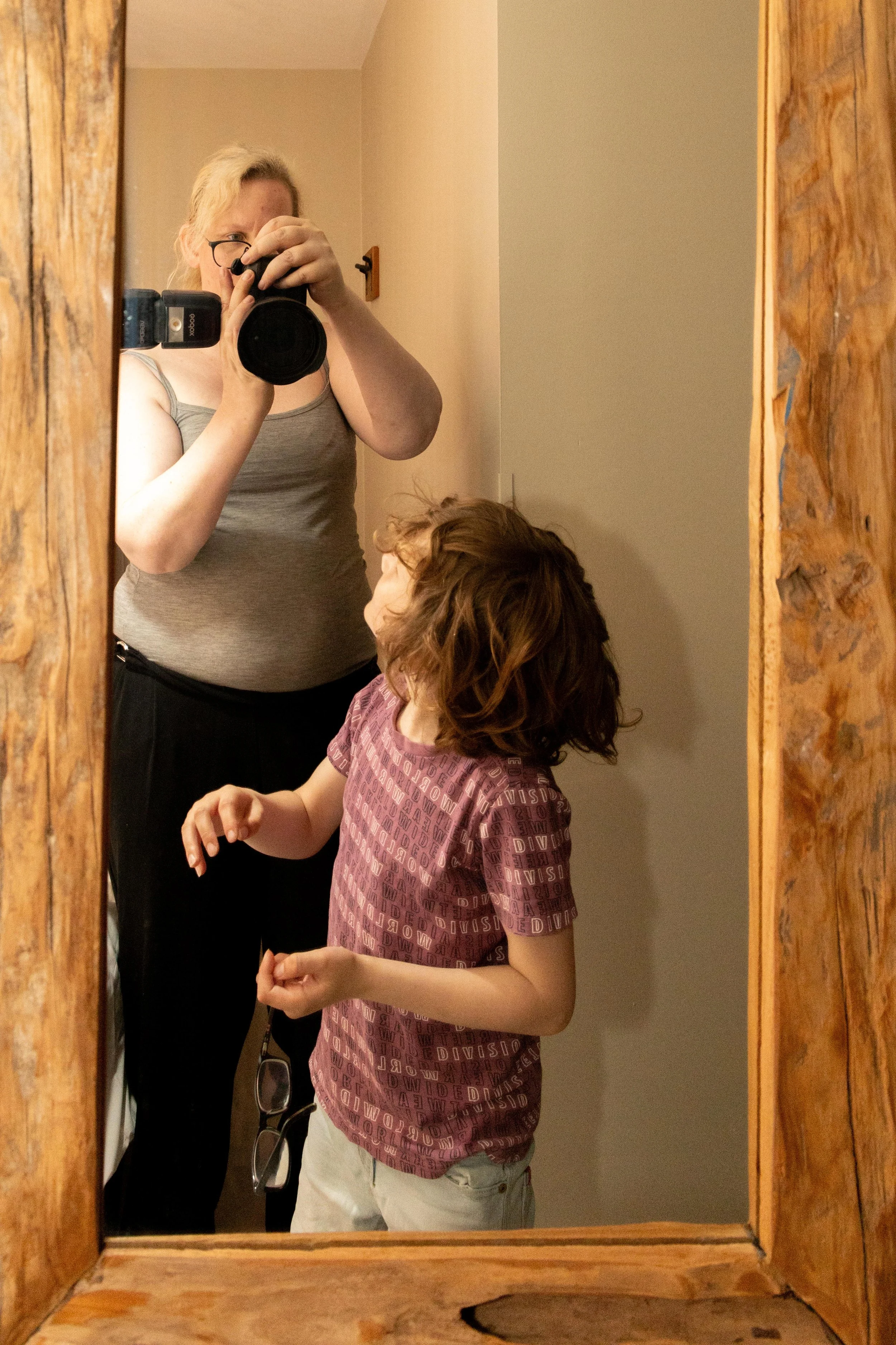 Een vrouw die selfies neemt met een camera in een houten frame spiegel, terwijl een kind naast haar staat.