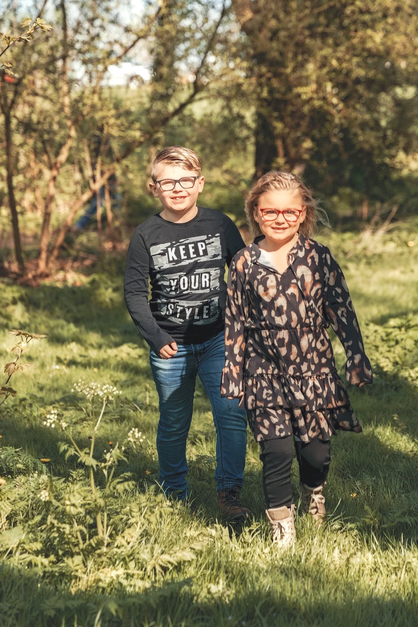 Two kids, a boy and a girl, walking in a green park with trees and grass, both smiling and wearing glasses on a sunny day.