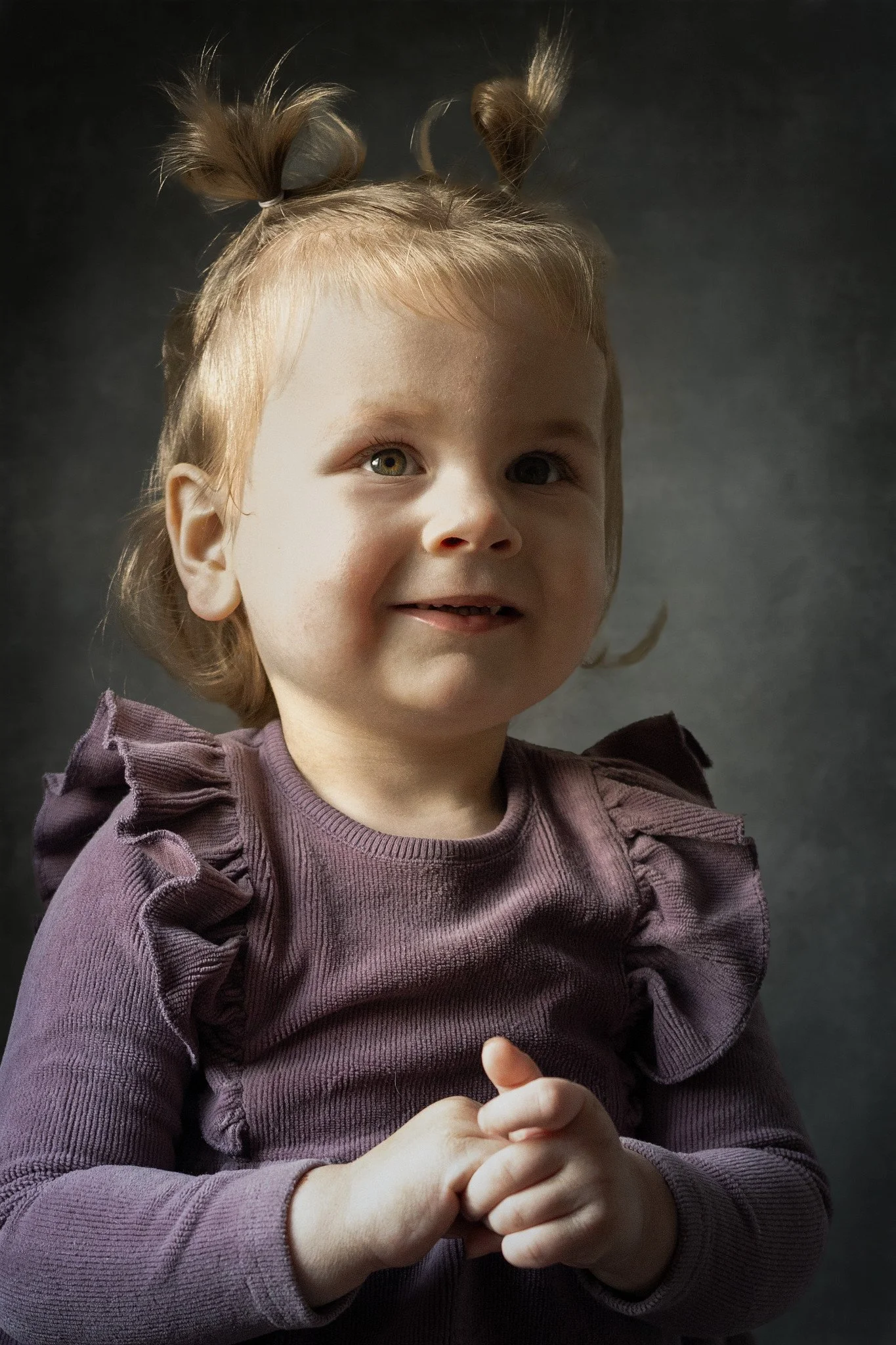 A young girl with blonde hair styled in two playful buns, wearing a mauve-colored sweater with ruffled shoulders, smiling softly against a dark background.