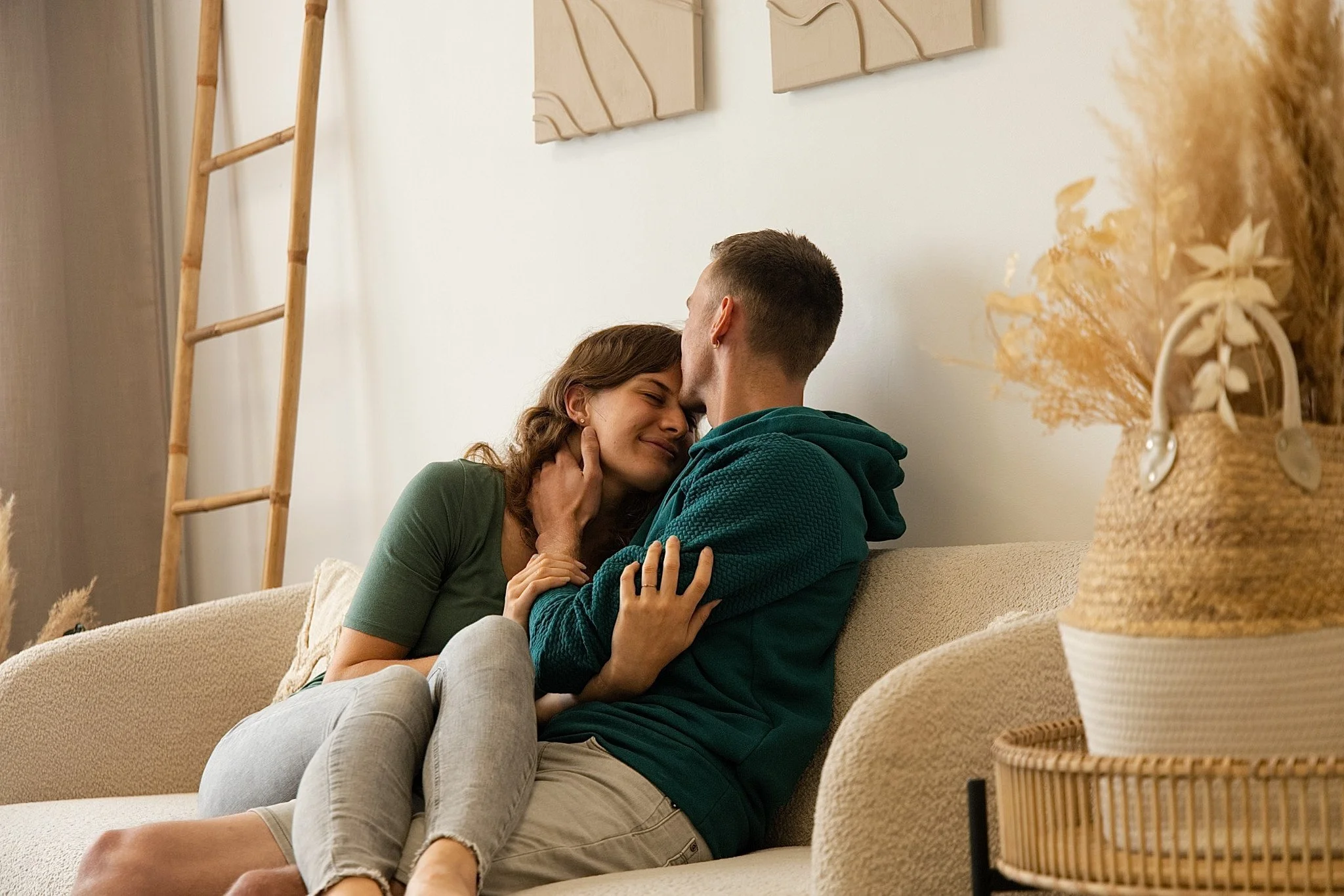 A couple sitting on a beige couch in a cozy living room, embracing and smiling at each other, with a wooden ladder and decorative art on the wall behind them, and a basket of dried flowers on a side table.