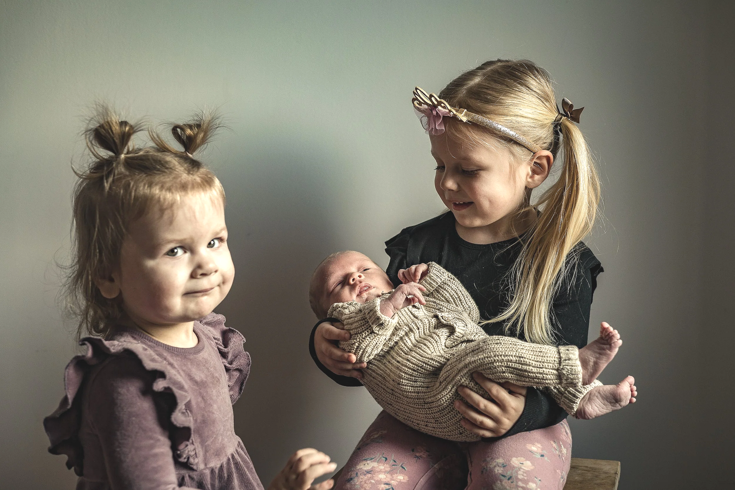 Two young girls, one with pigtails and purple dress, and the other with long blonde hair and butterfly headband, holding a newborn baby in knitted clothing, against a neutral background.