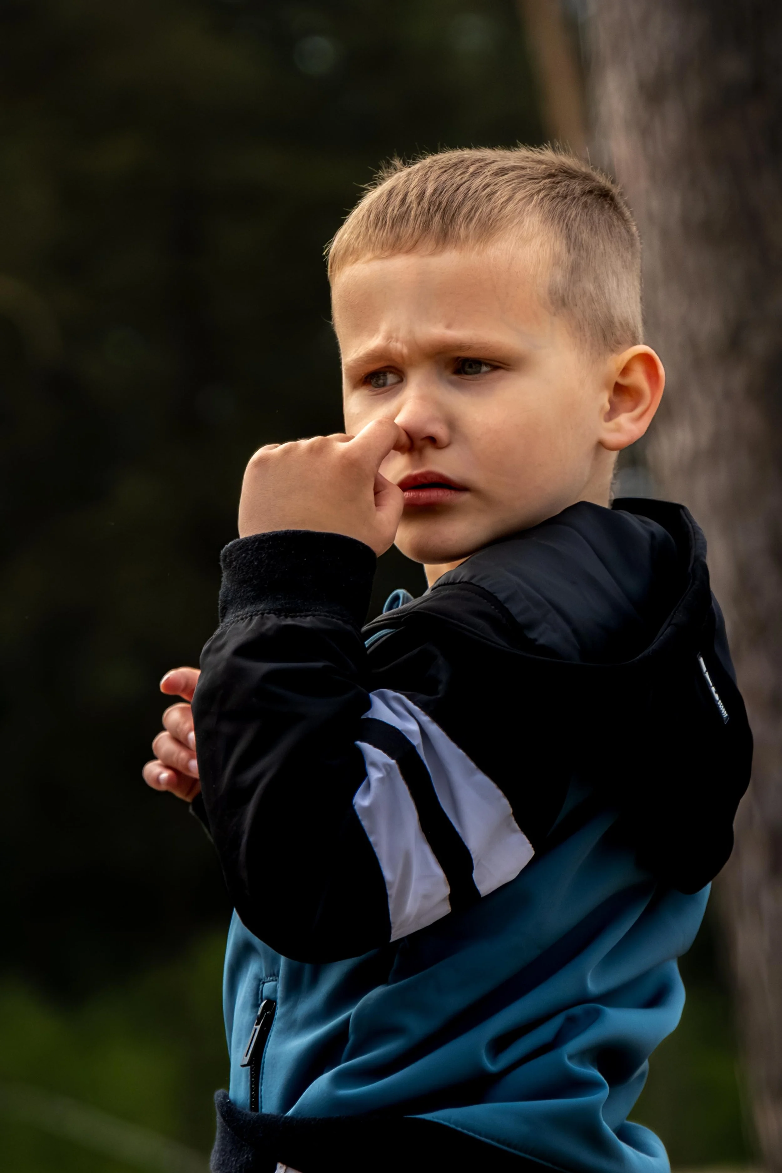 Tienerjongen in een blauw en zwart jack met zijn hand in neus, gericht naar de rechterkant.