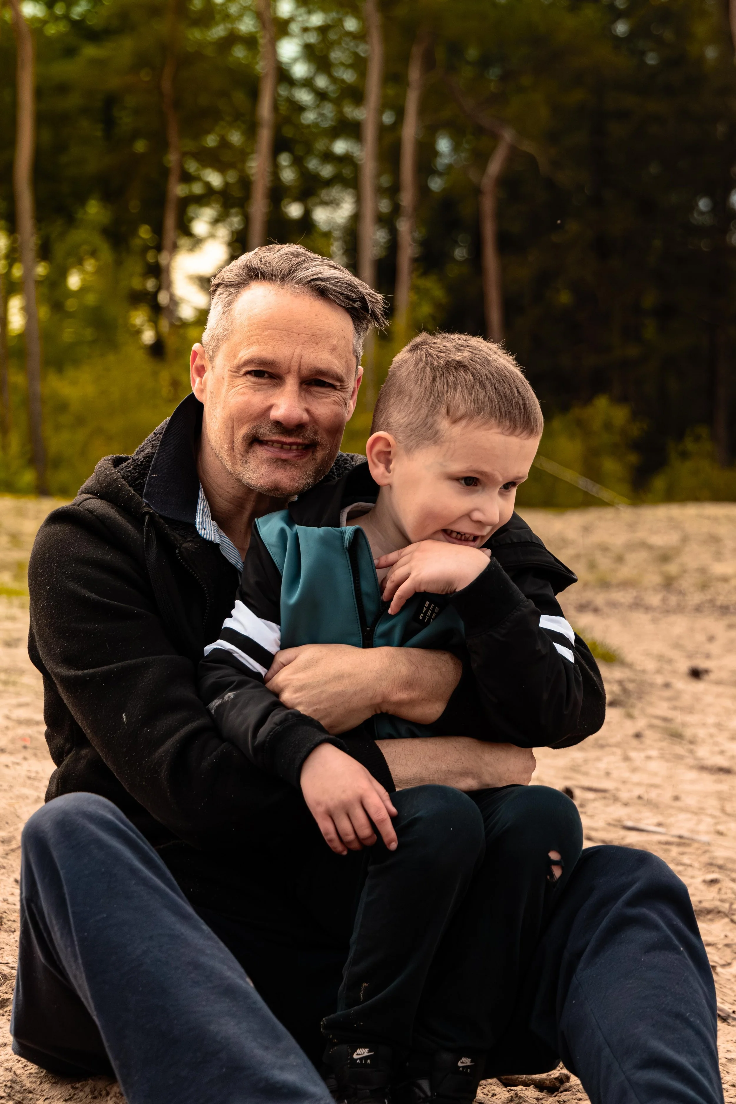 Een oudere man met grijs haar en een jong kind zitten samen buiten op het zand, omringd door bomen, en genieten van elkaars gezelschap.