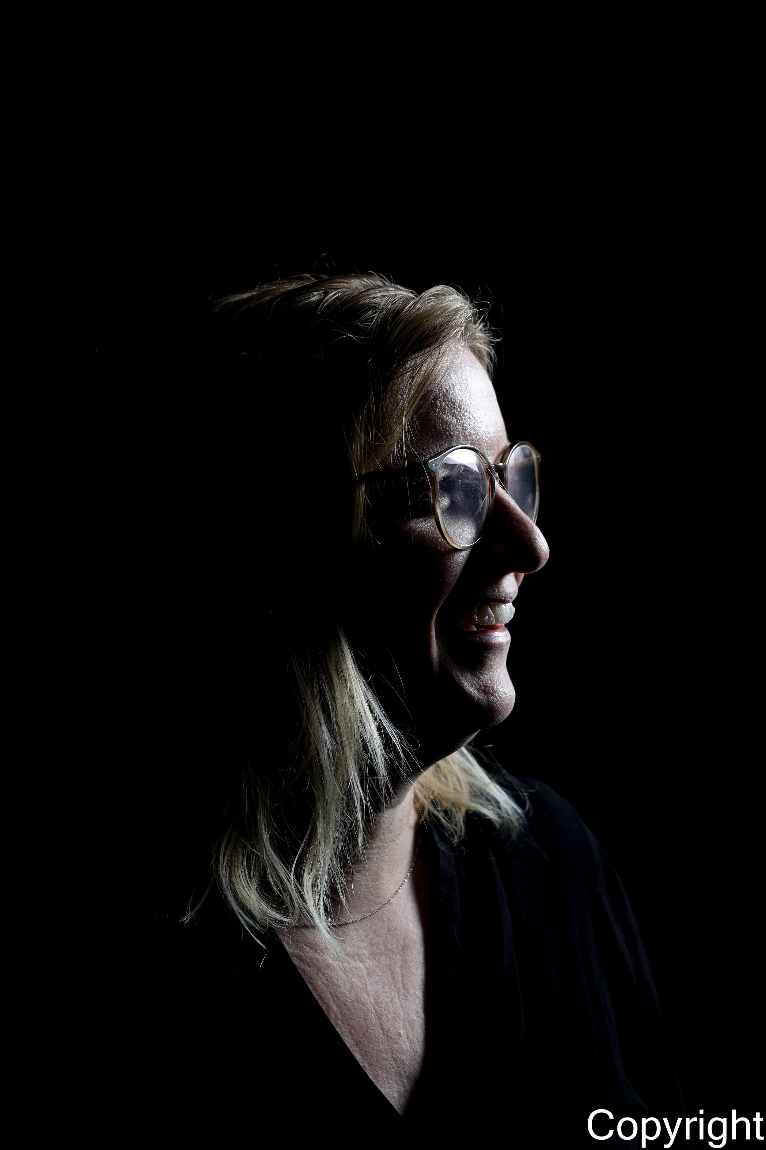 Close-up van een lachende vrouw met blond haar en bril, kijkend naar rechts, tegen een zwarte achtergrond.
