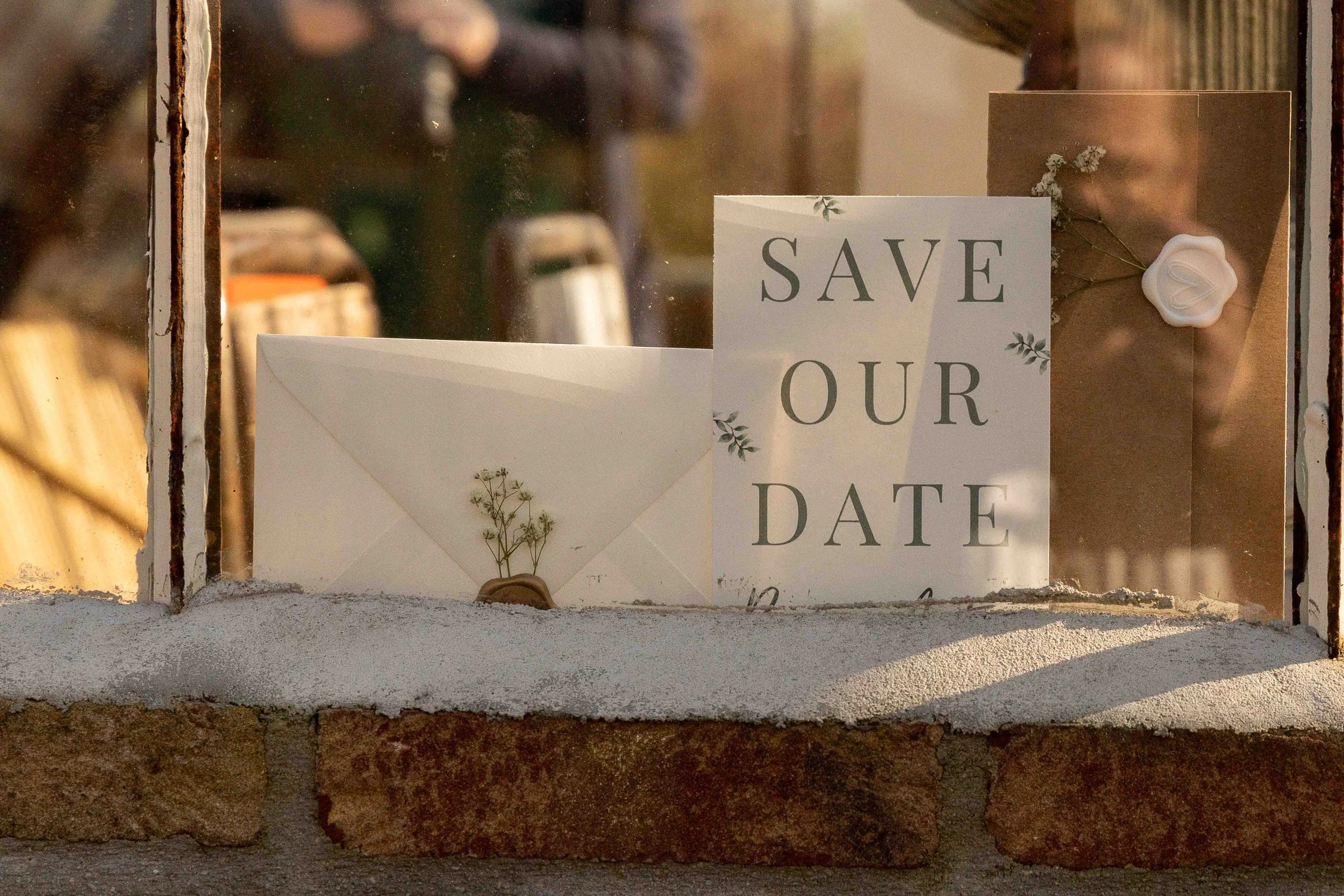 Een raam met een witte kaart die zegt 'Save Our Date' en een envelop met bloemen. Naast de kaart een waxinelichtje en bloemen onder een waxinelichtje.