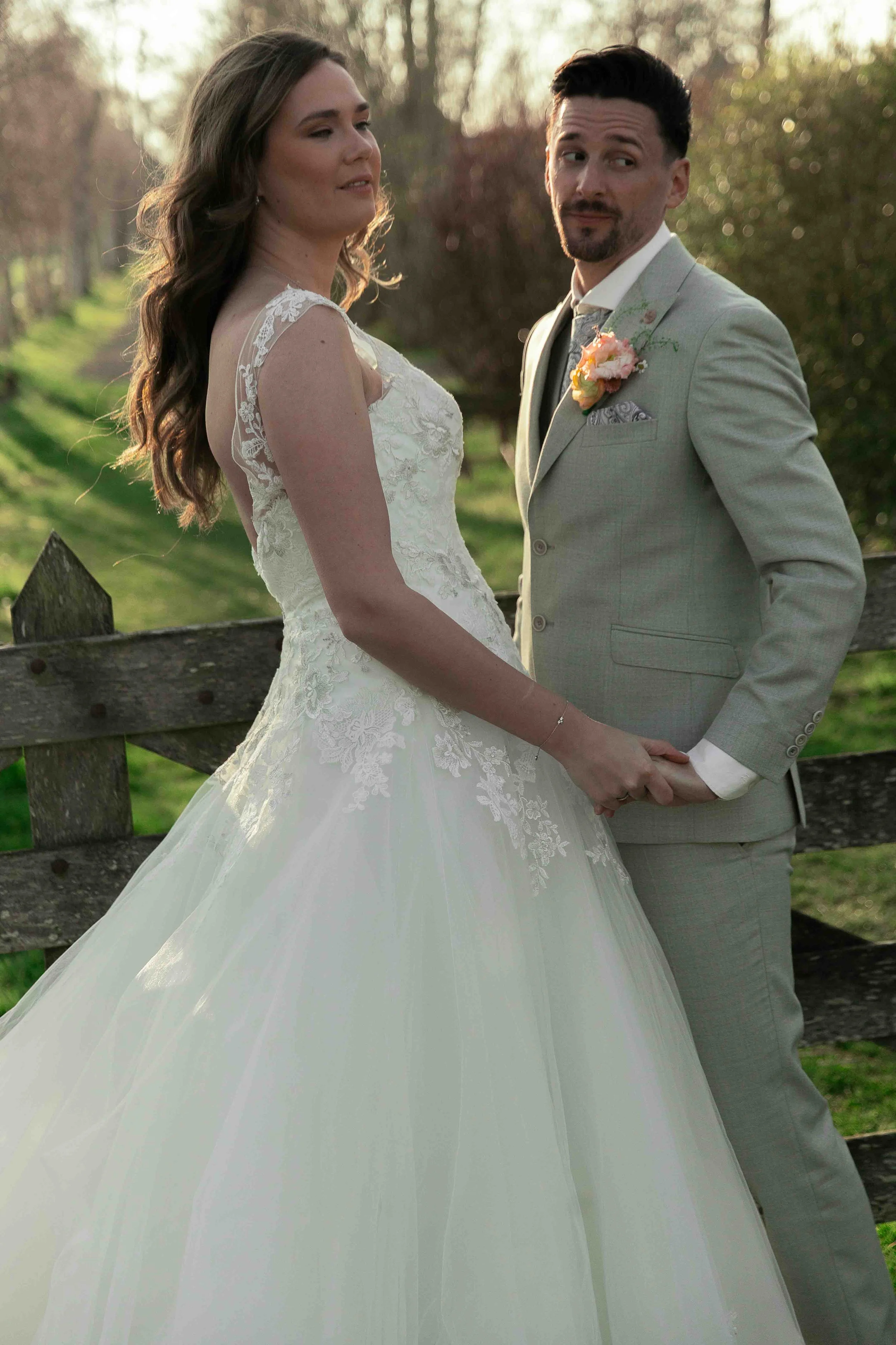 Een bruiloftsfoto van een stel dat hand in hand staat in een park, de vrouw draagt een witte trouwjurk en de man een lichtgrijze pak met een bloemenbroche.