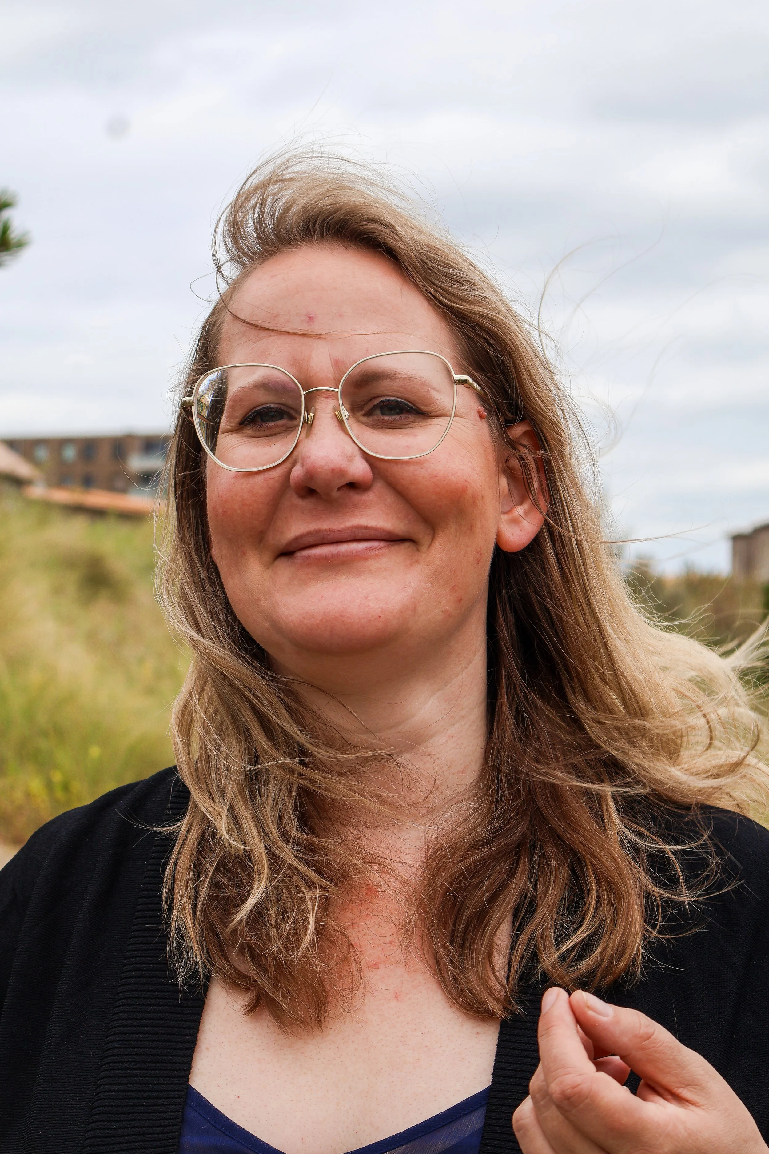 A woman with glasses smiling outdoors with grass and buildings in the background.