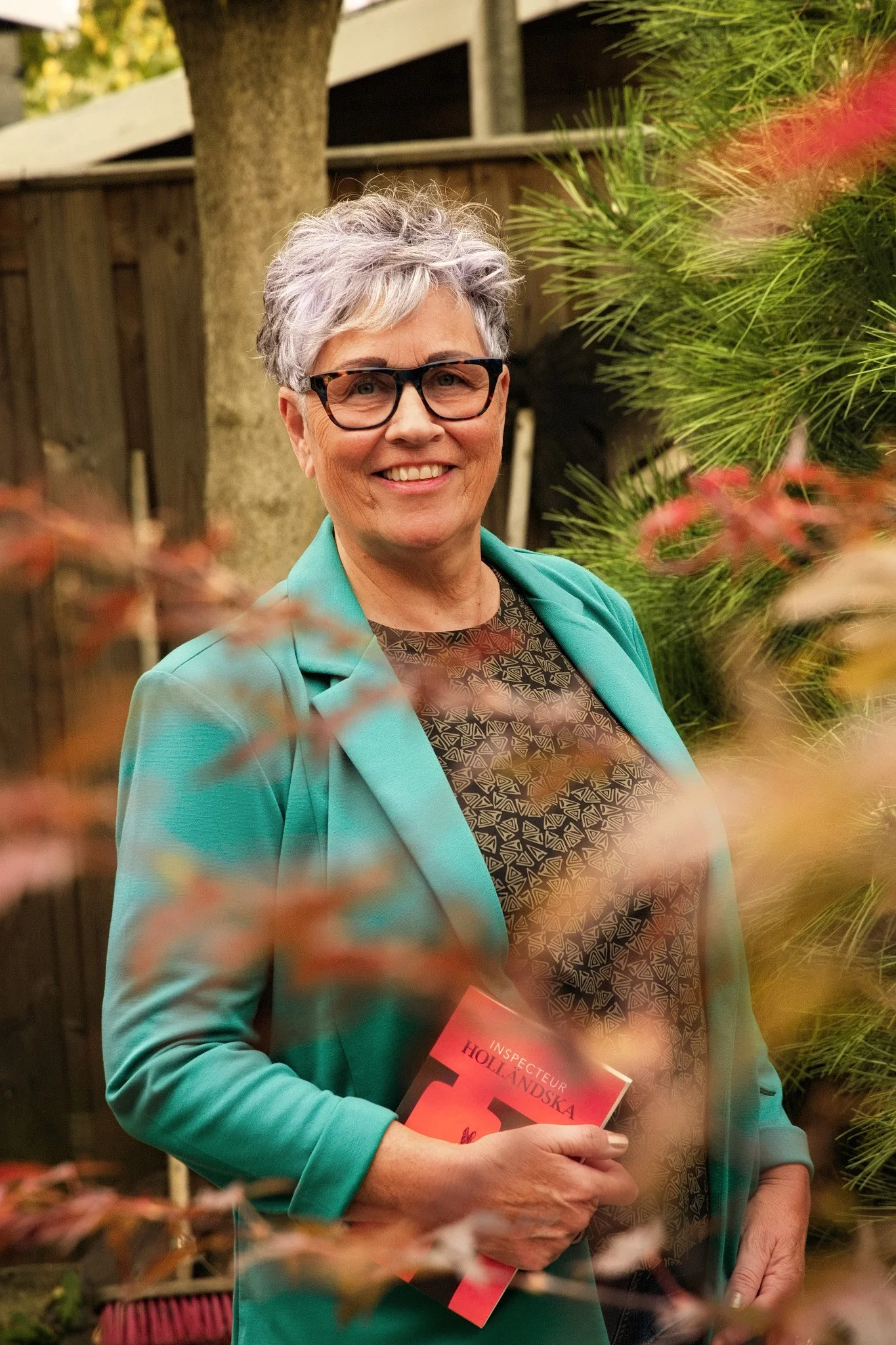 An older woman with short gray hair and glasses, smiling, standing outdoors behind greenery and a wooden fence, holding a red book titled 'Inspecteur Hollandska'.