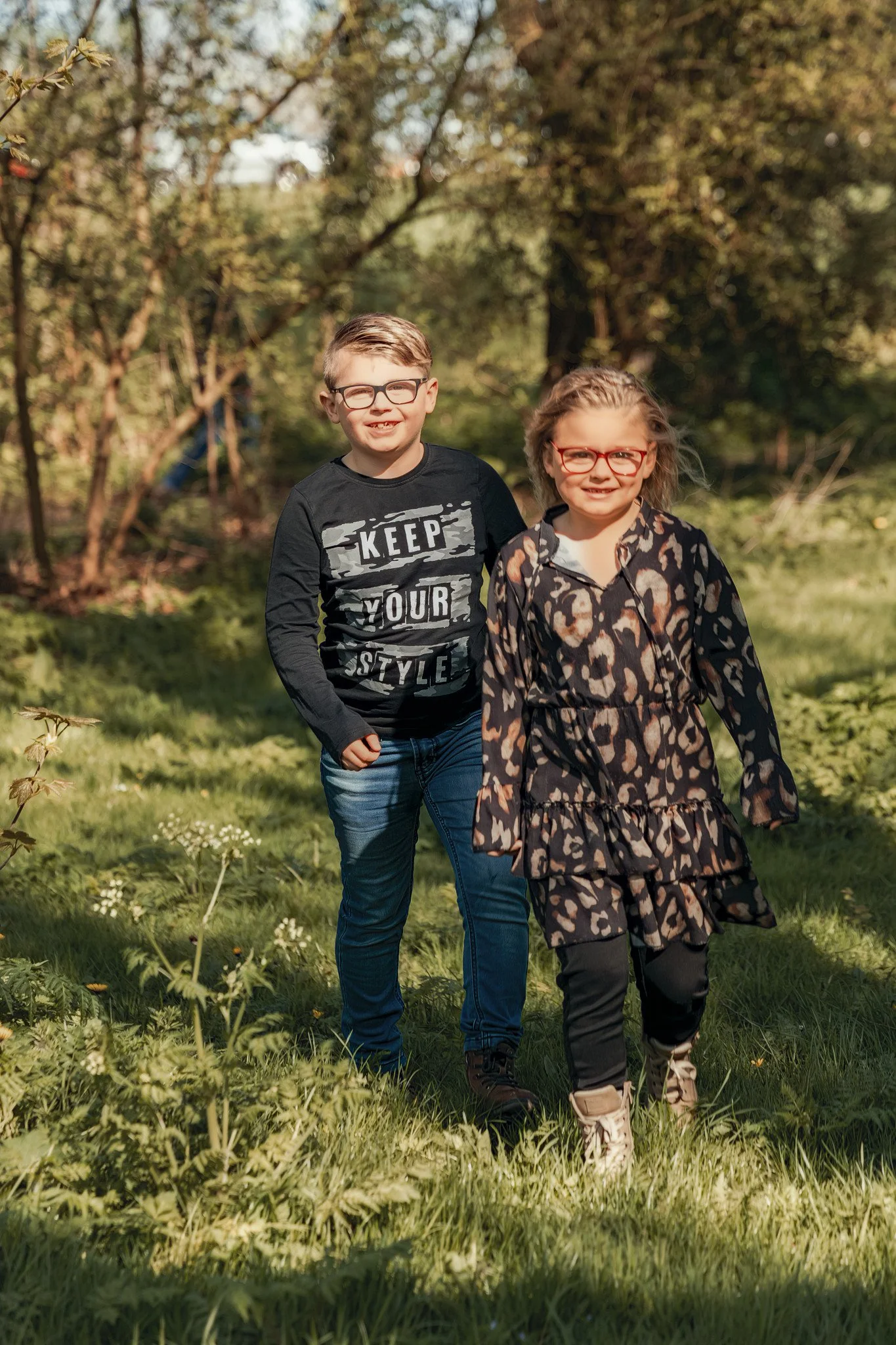 Two children, a boy and a girl, walking through a grassy park on a sunny day, both wearing glasses and casual clothing, smiling.