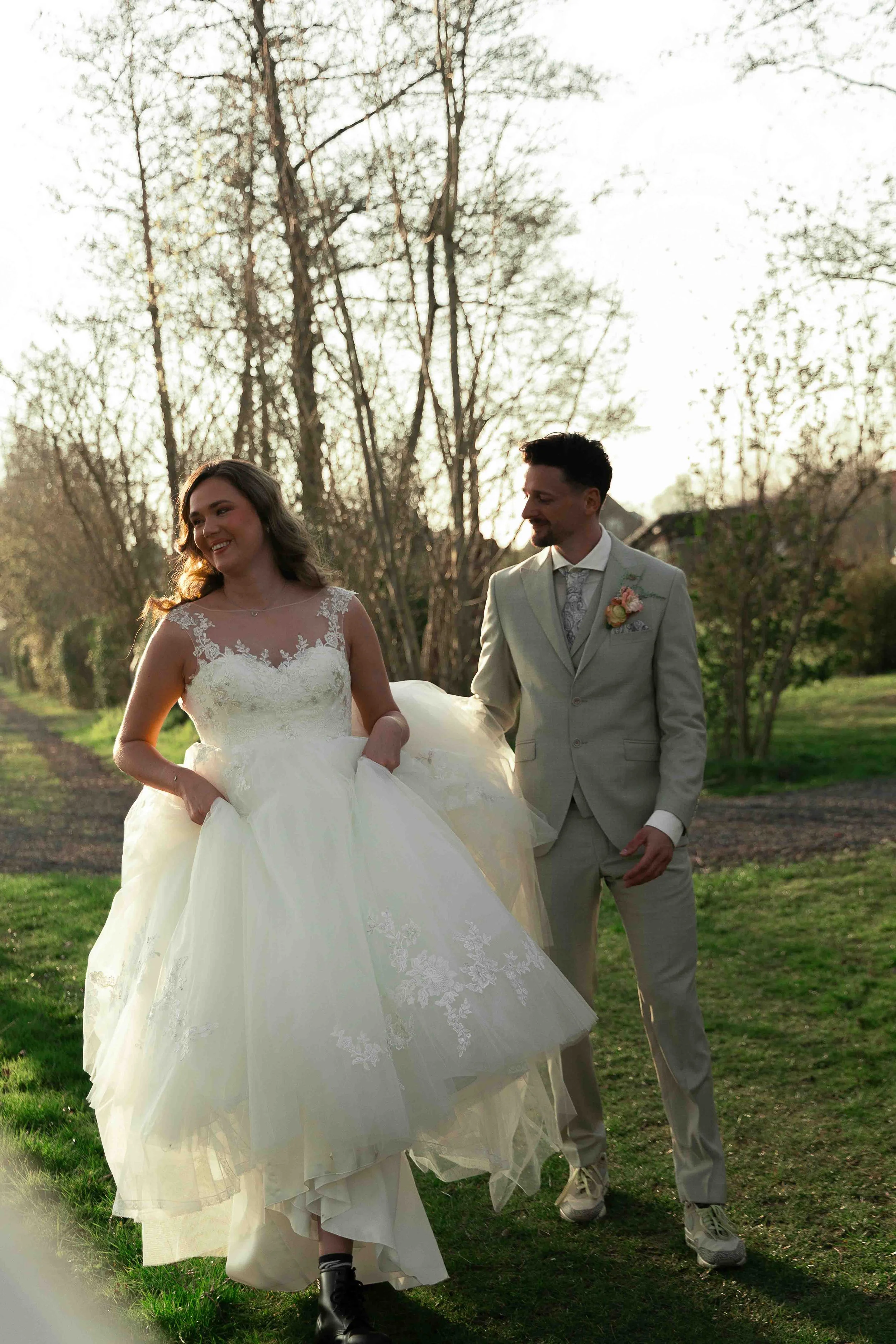Een bruid en bruidegom lopen samen in een tuin, de bruid draagt een witte bruidsjurk en laarzen, de bruidegom draagt een grijze pak met boutonnière, het is avond en de bomen in de achtergrond zijn kaal.