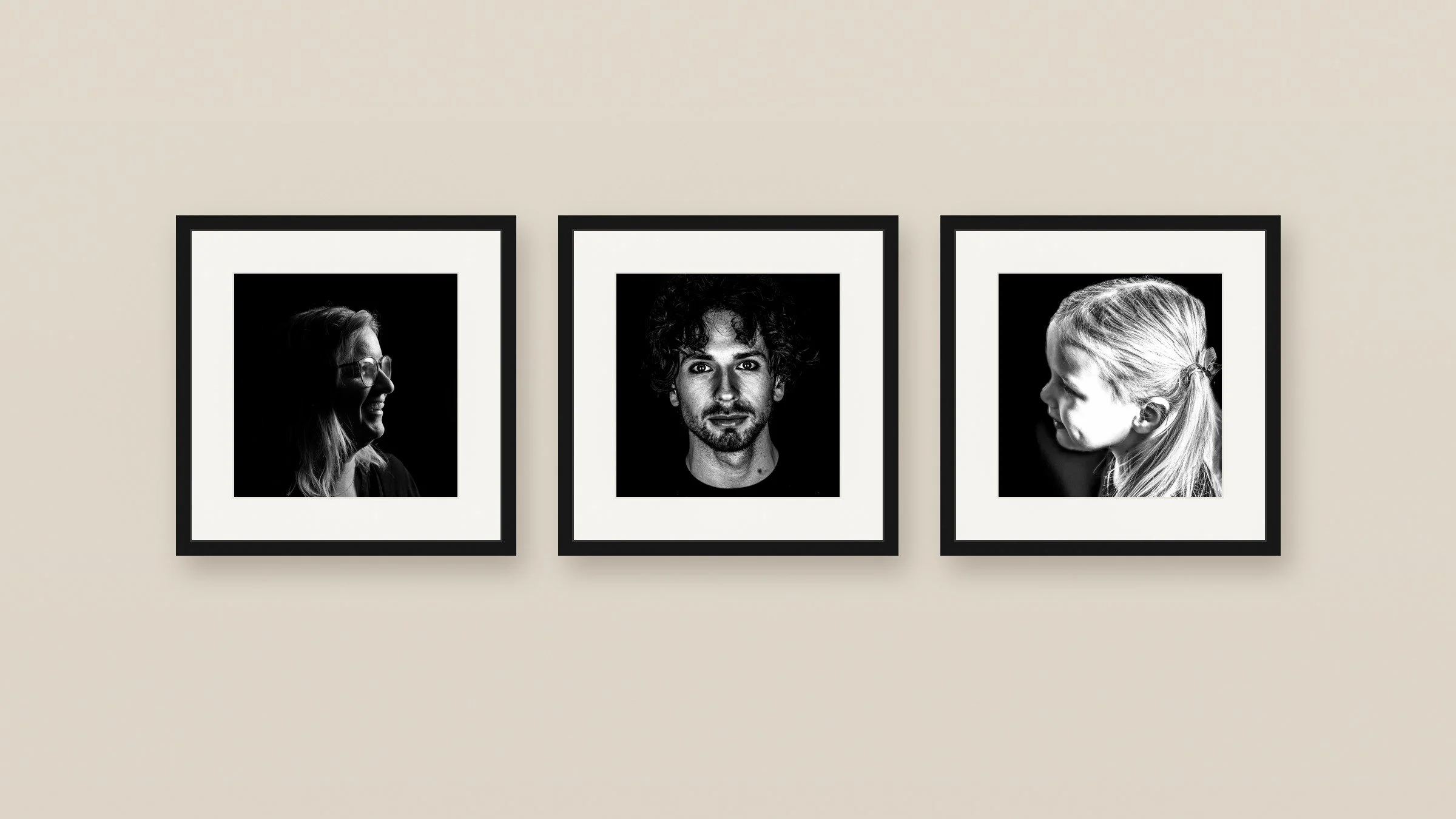 Drie zwart-wit fotoafdrukken van mensen in lijstformaten hangen aan een beige muur. De links twee vrouwen in profiel, de middelste man direct voor de camera met een neutrale uitdrukking.