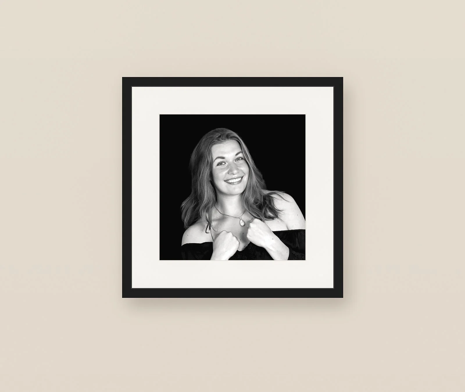 Zwart-wit portretfoto van een jonge vrouw met glimlach, gekleed in een off-shoulder top en sieraden, tegen een zwarte achtergrond in een wit frame.