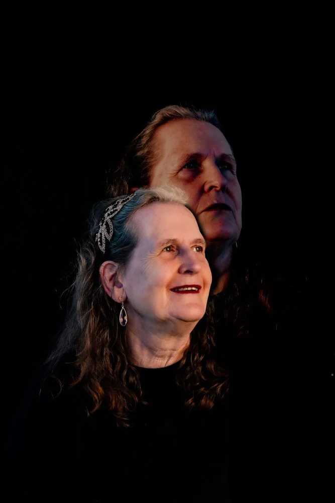 Twee vrouwen, een oudere vrouw met bruin haar en een jonge vrouw met licht haar en een haarband, kijken omhoog tegen een zwarte achtergrond.